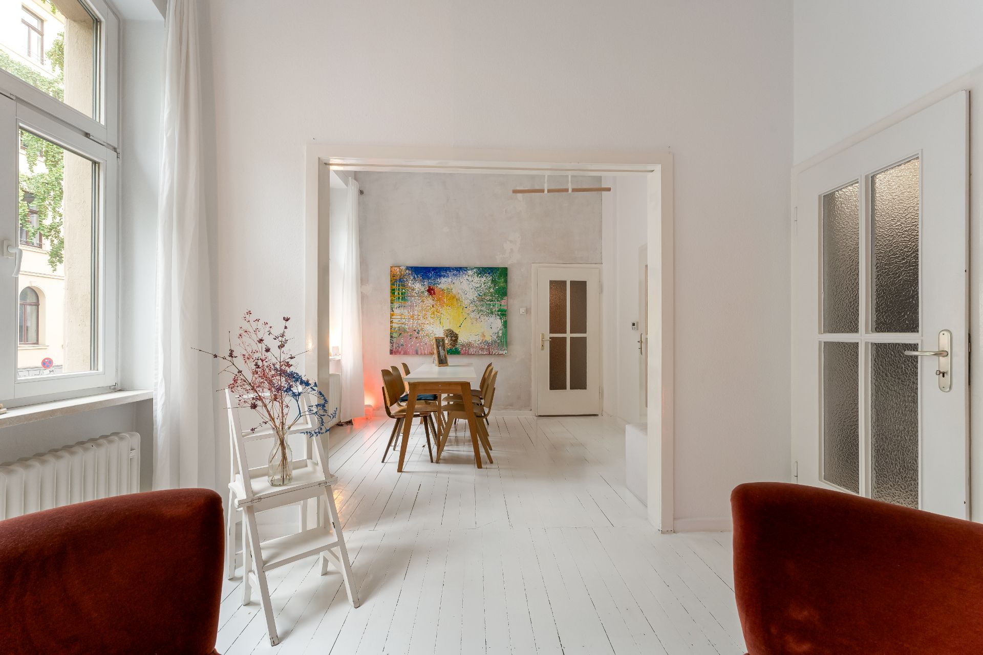 Helles Wohnzimmer mit weißen Wänden, Holztisch, Stühlen und buntem Gemälde an der Wand.