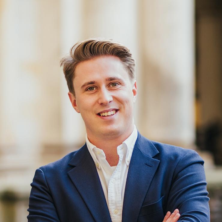 Junger Mann im blauen Sakko steht mit verschränkten Armen vor unscharfem Hintergrund.