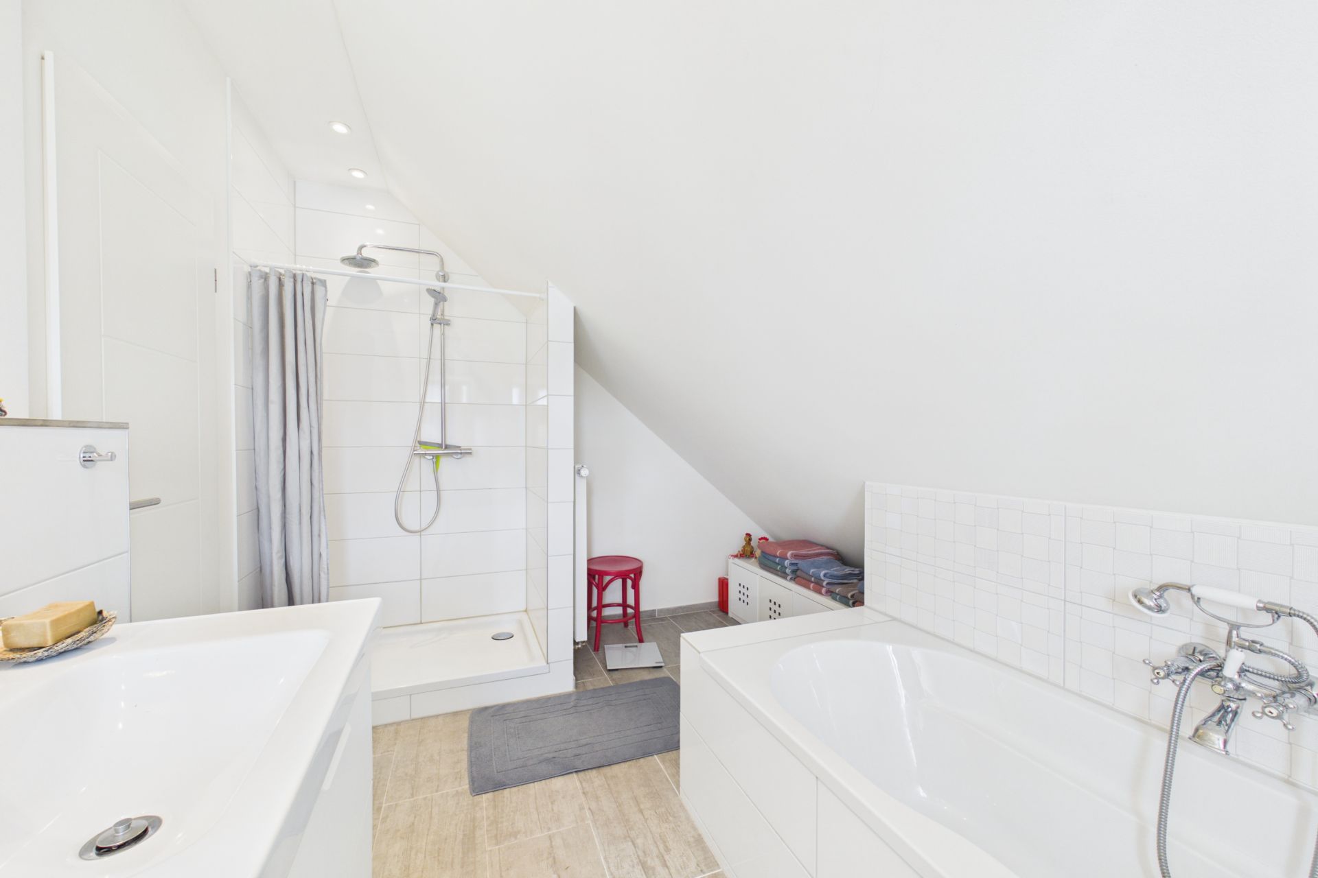 Helles Badezimmer mit Dusche, Badewanne, Waschbecken und rotem Hocker unter einer Dachschräge.
