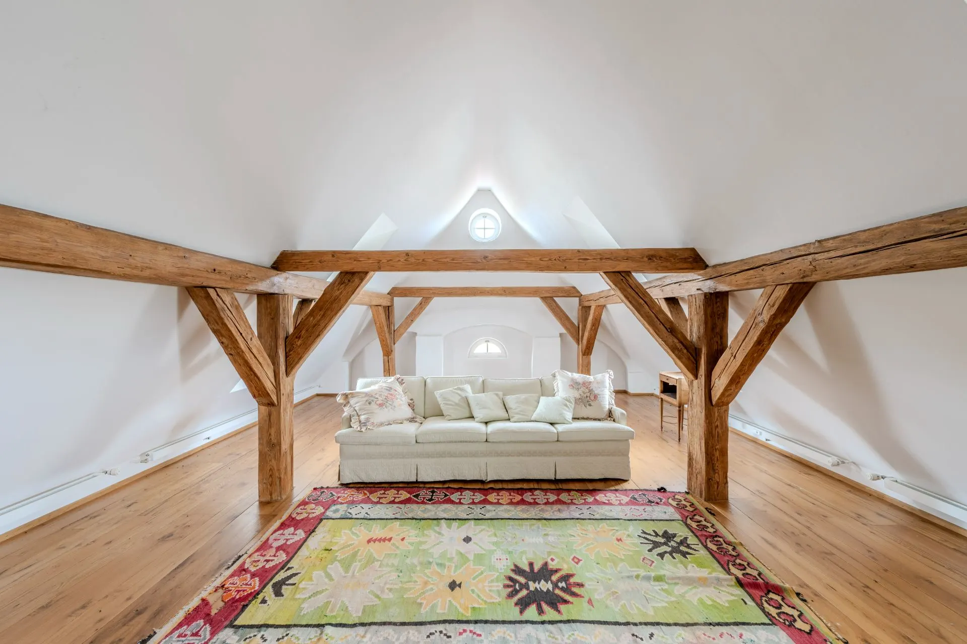 Helles Dachzimmer mit Holzbalken, Sofa und buntem Teppich auf Holzboden.
