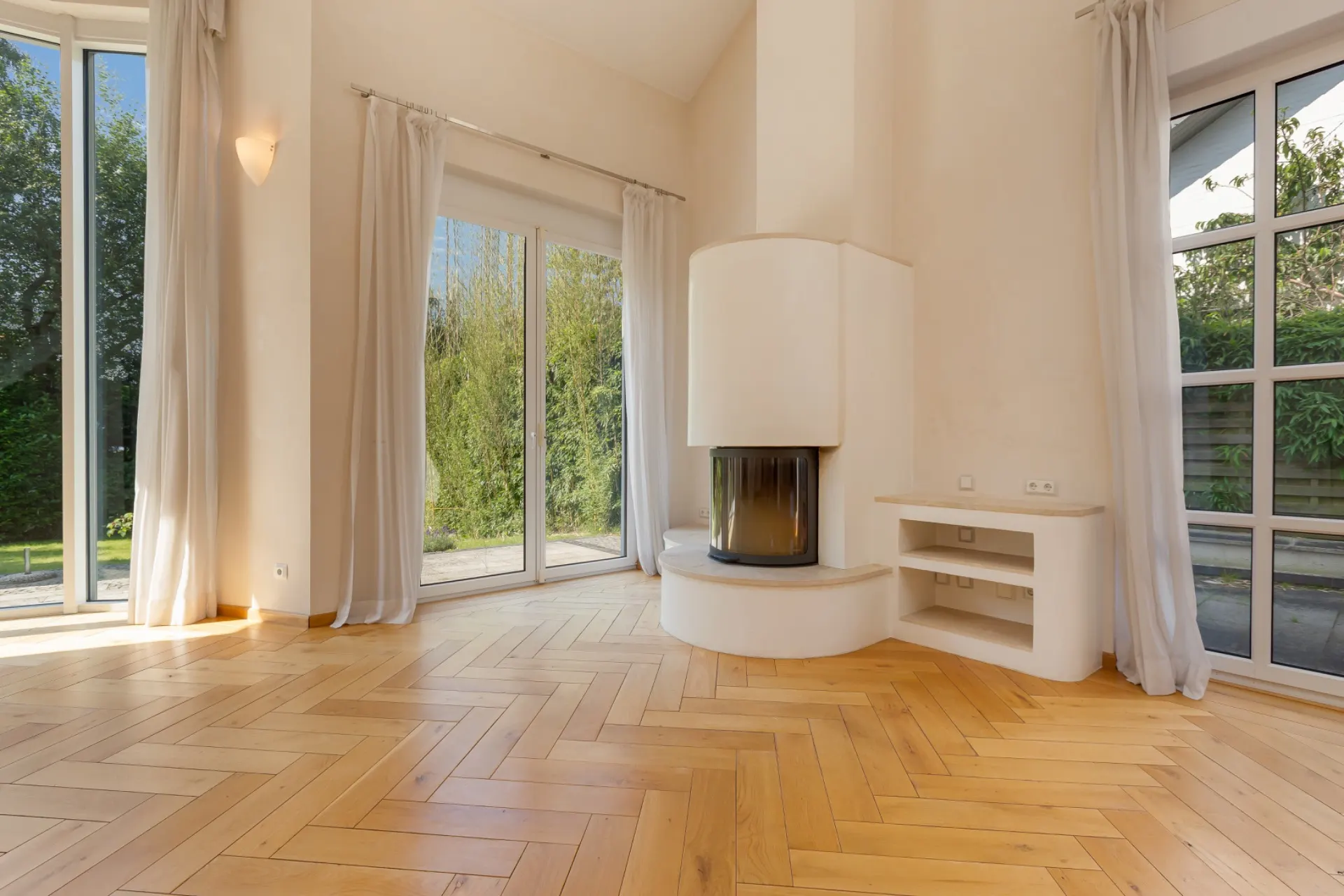 Helles Wohnzimmer mit Kamin, Parkettboden und großen Fenstern mit Vorhängen.