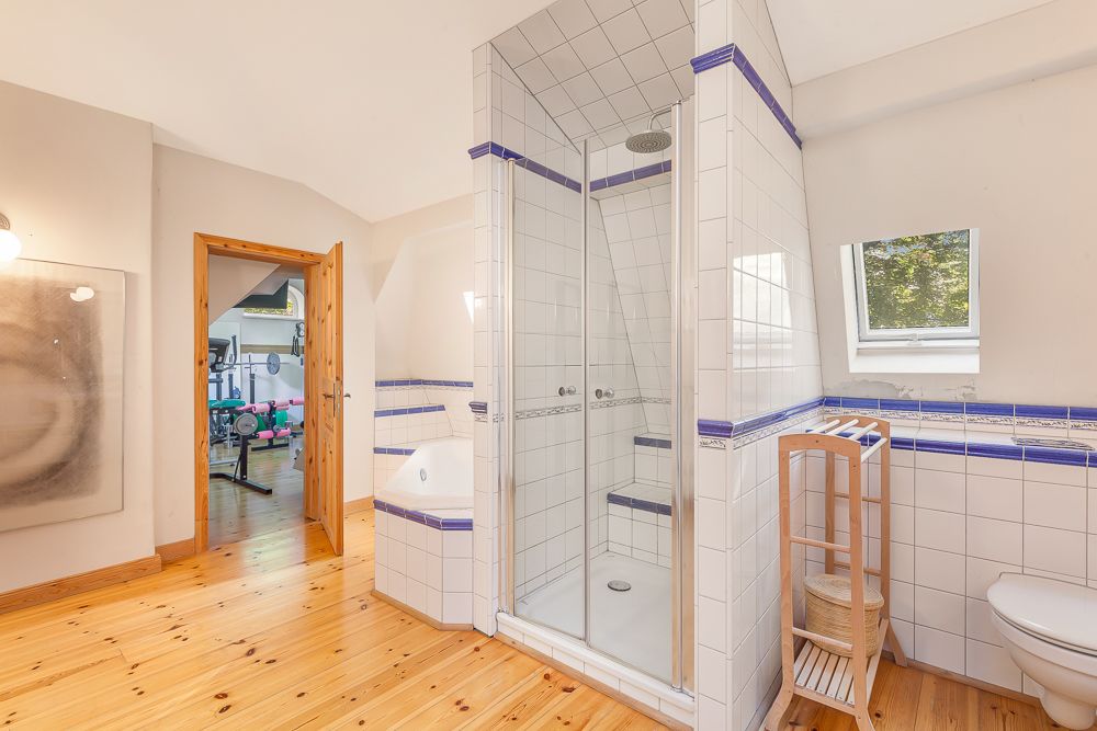 Helles Badezimmer mit Dusche, Fenster, Toilettenpapierhalter und Blick in einen Fitnessraum.