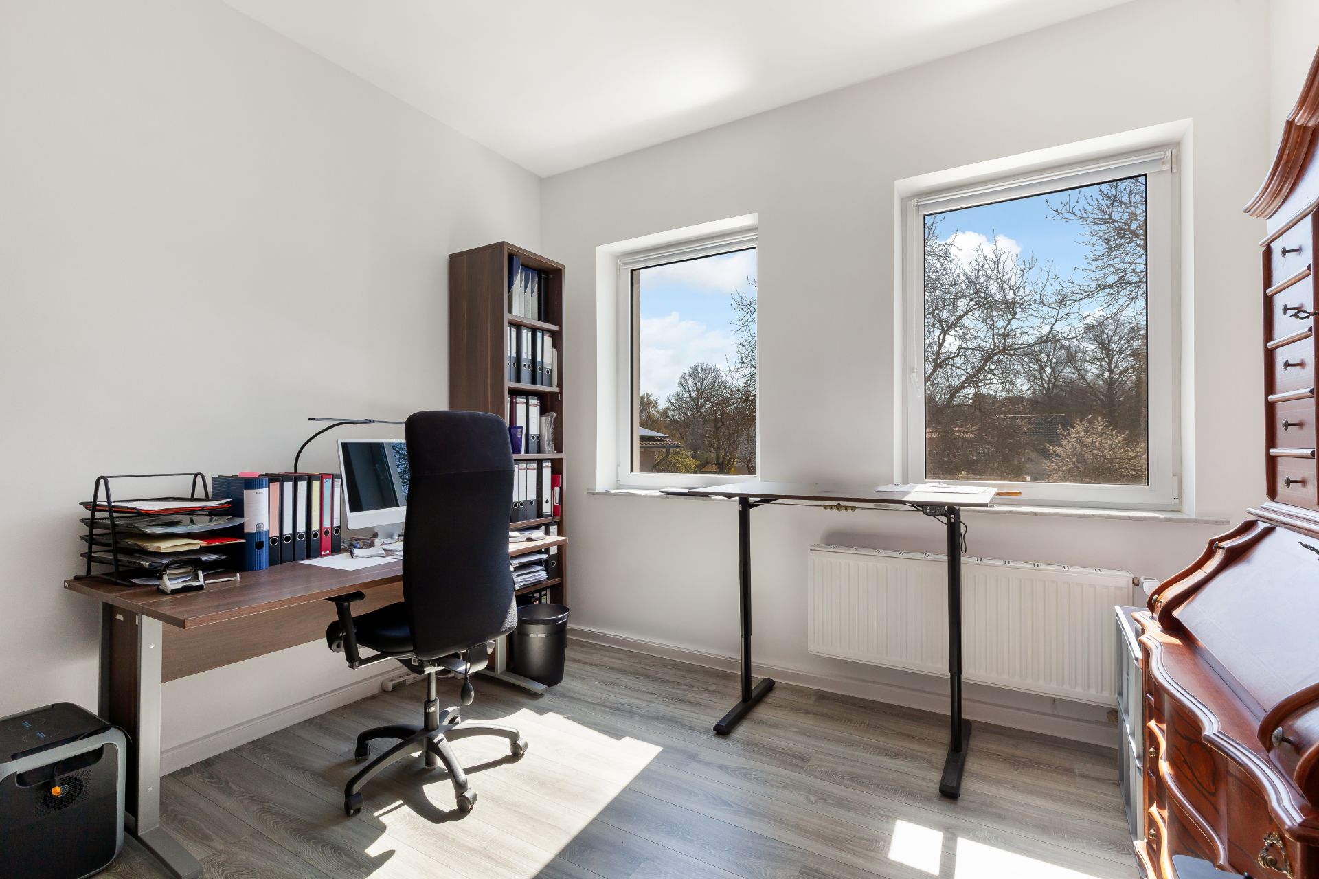 Helles Arbeitszimmer mit Schreibtisch, Computer, Regal und zwei großen Fenstern.
