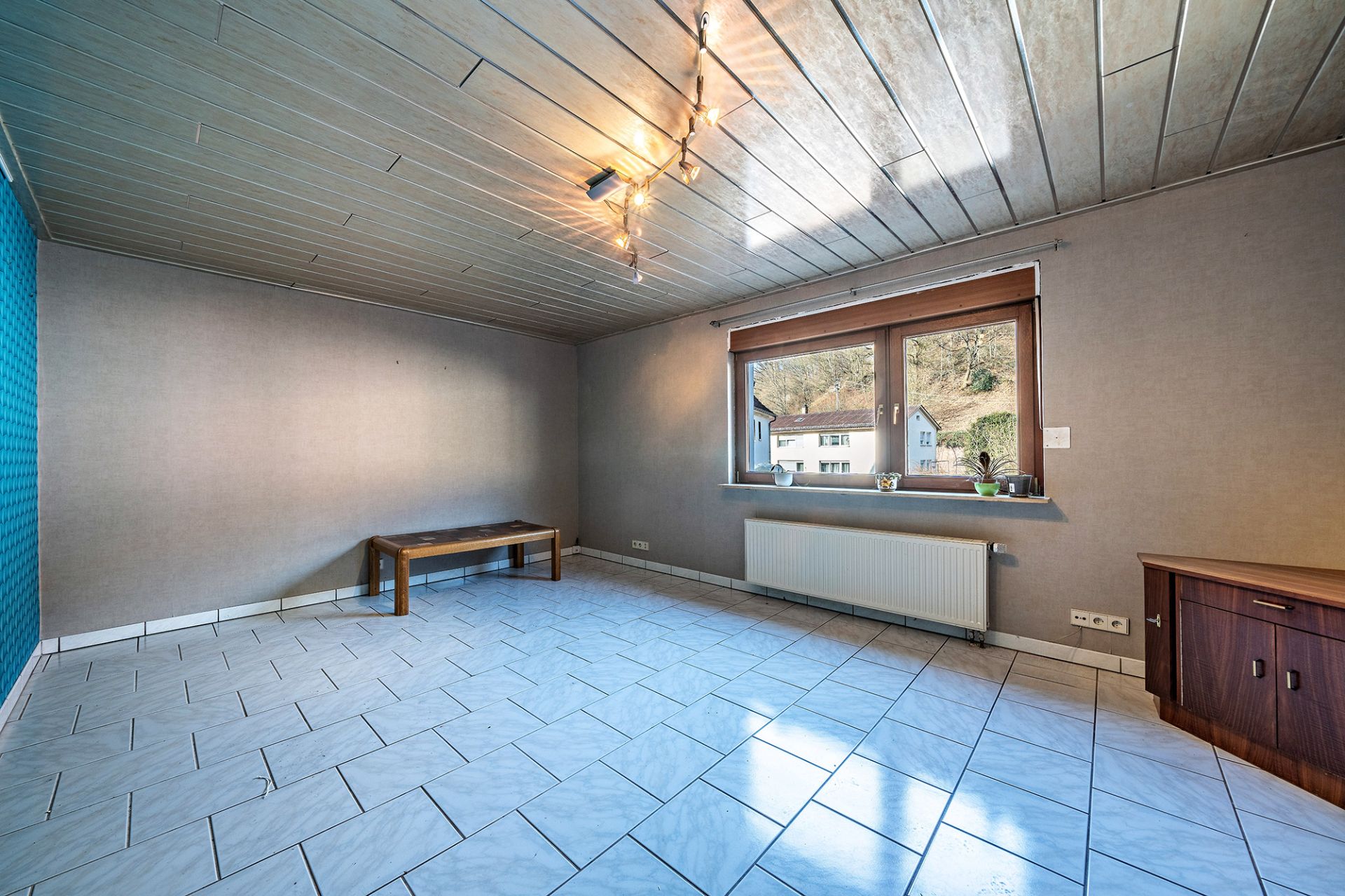 Leeres Wohnzimmer mit weißen Fliesen, Holzdecke, großem Fenster und kleinem Holztisch.