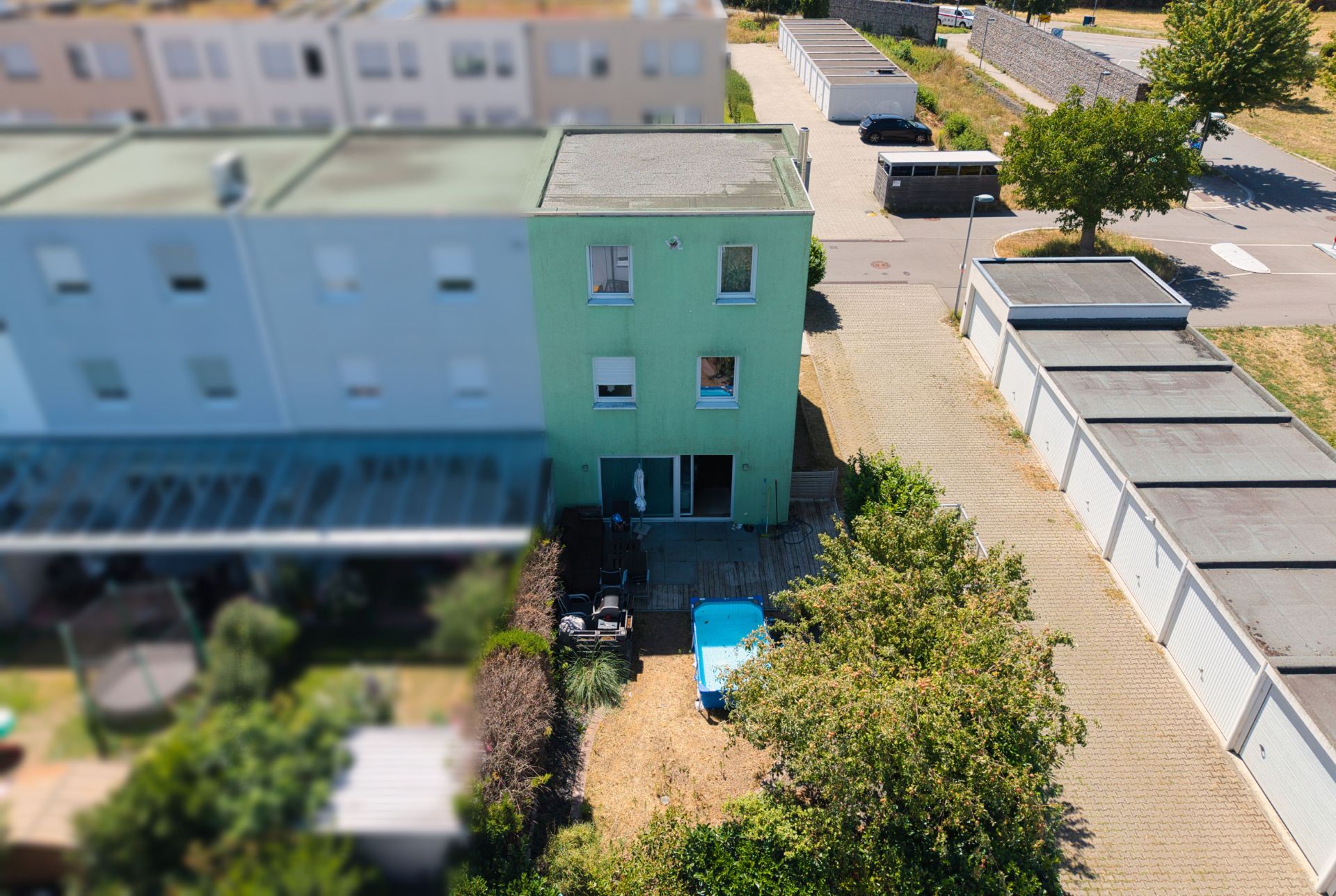 Grünes Reihenhaus mit Garten, Terrasse und Parkplatz, daneben mehrere Garagen und ein Baum.