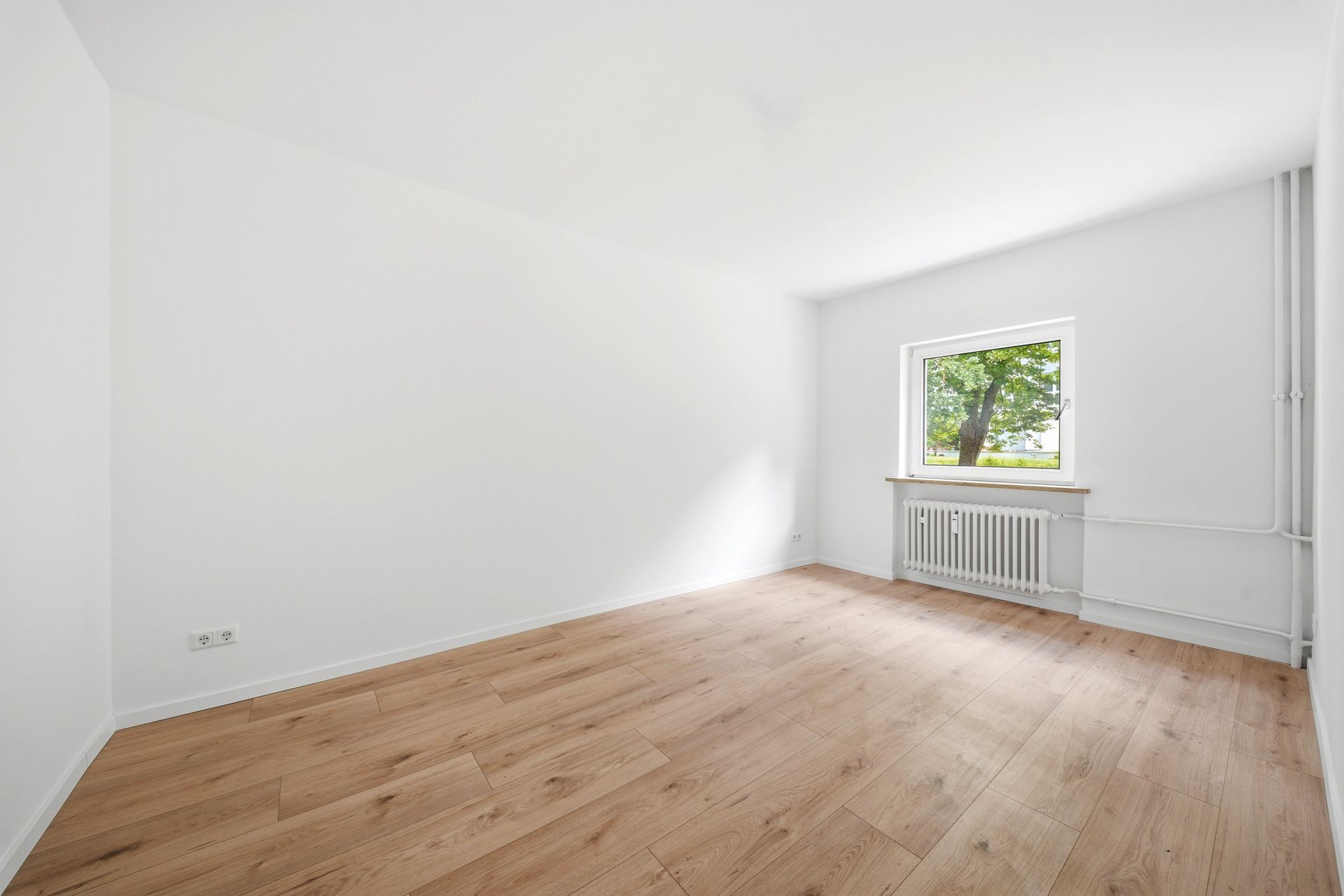 Leerer Raum mit Holzfußboden, weiß gestrichenen Wänden und einem Fenster mit Blick auf Bäume.