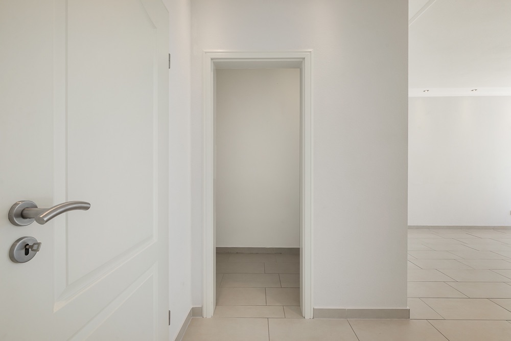 Weißer Flur mit offener Tür und gefliestem Boden in einer Wohnung.