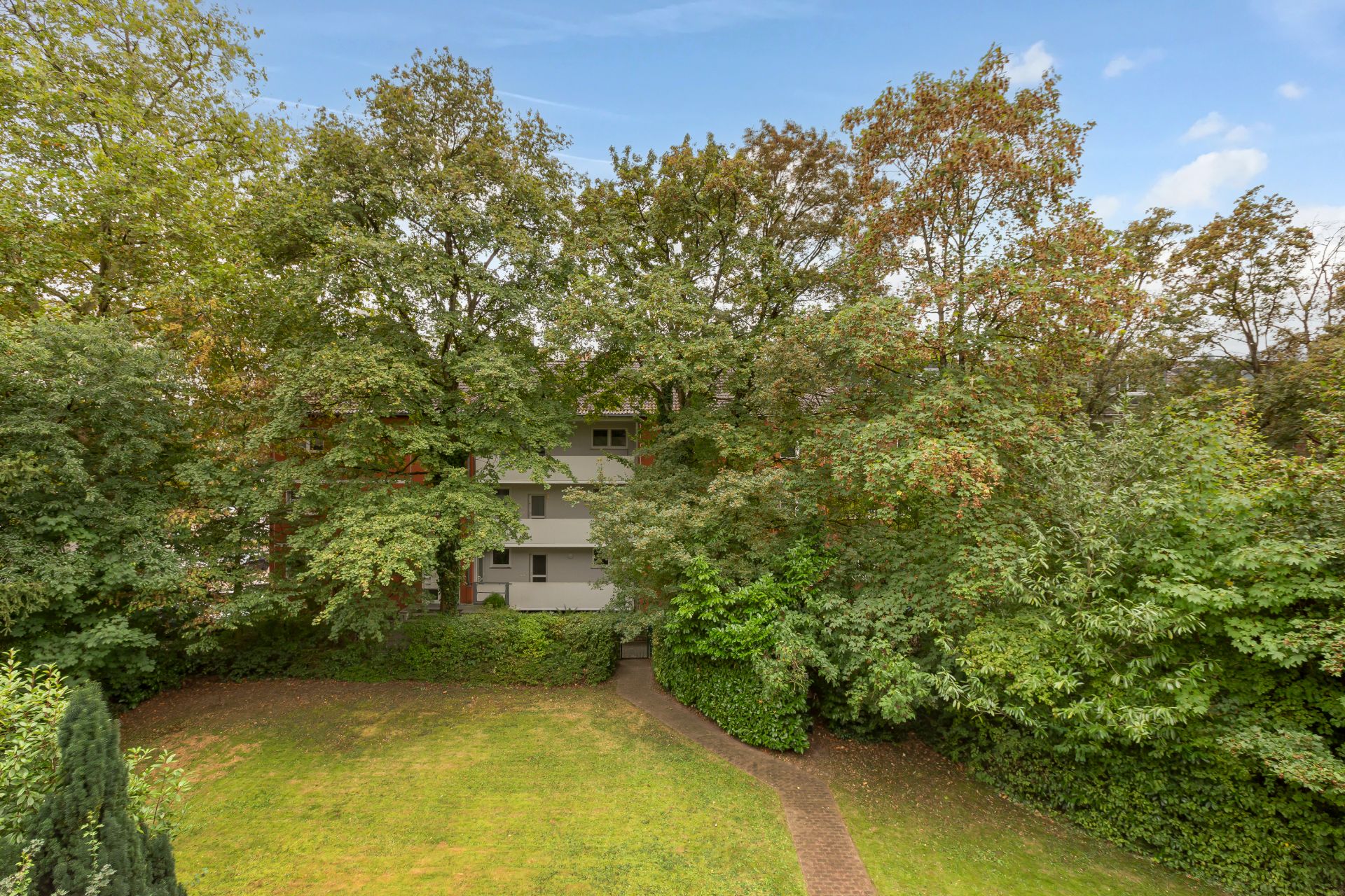 Wohnhaus hinter Bäumen, davor Garten mit Wiese und Gehweg.