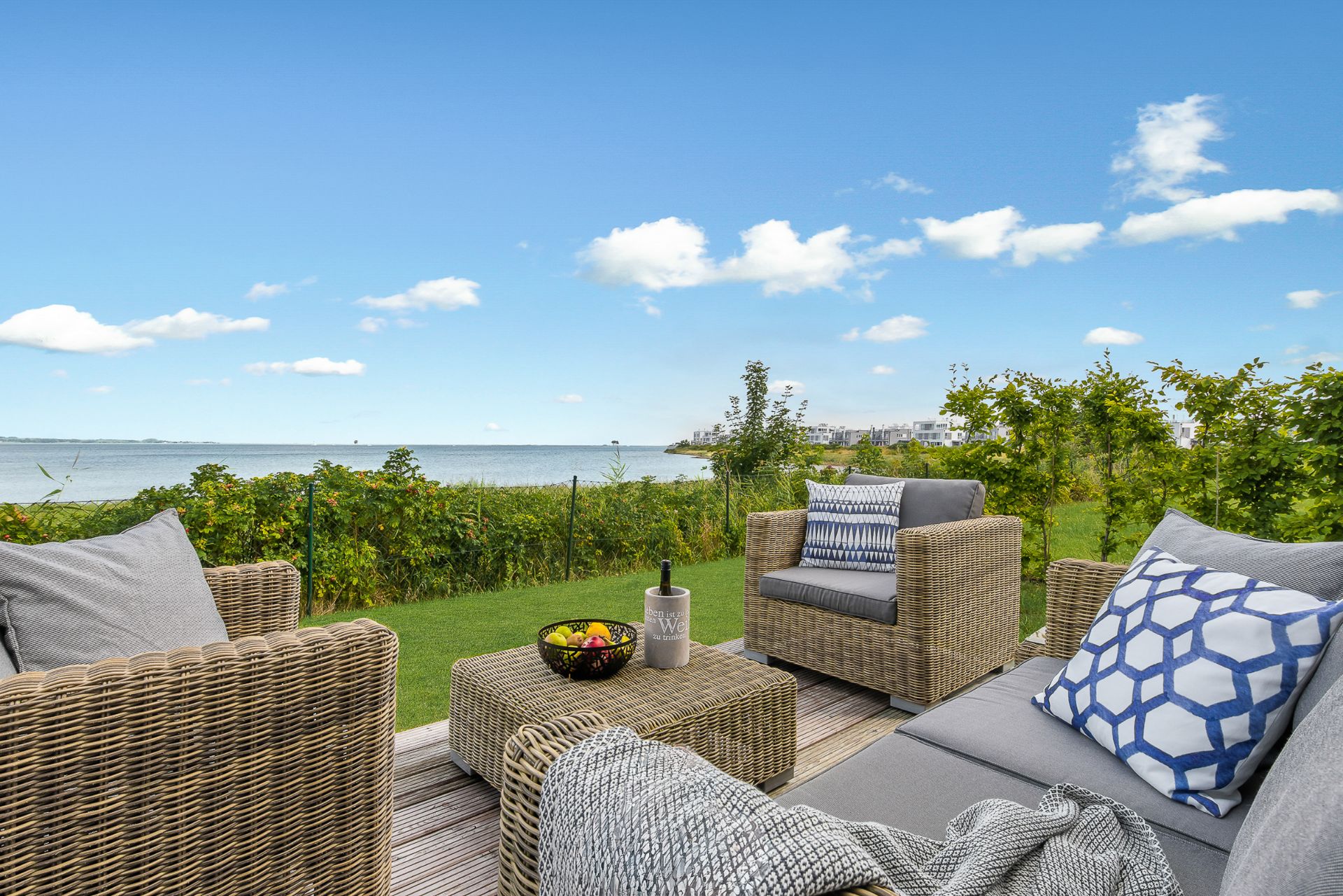 Terrasse mit Rattanmöbeln, Kissen und Tisch, Blick auf Garten und Wasser im Hintergrund.