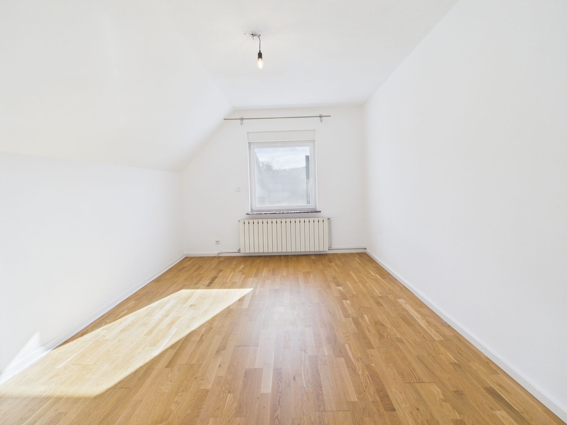 Helles, leeres Zimmer mit weiß gestrichenen Wänden, Holzfußboden und einem Fenster mit Heizkörper darunter.
