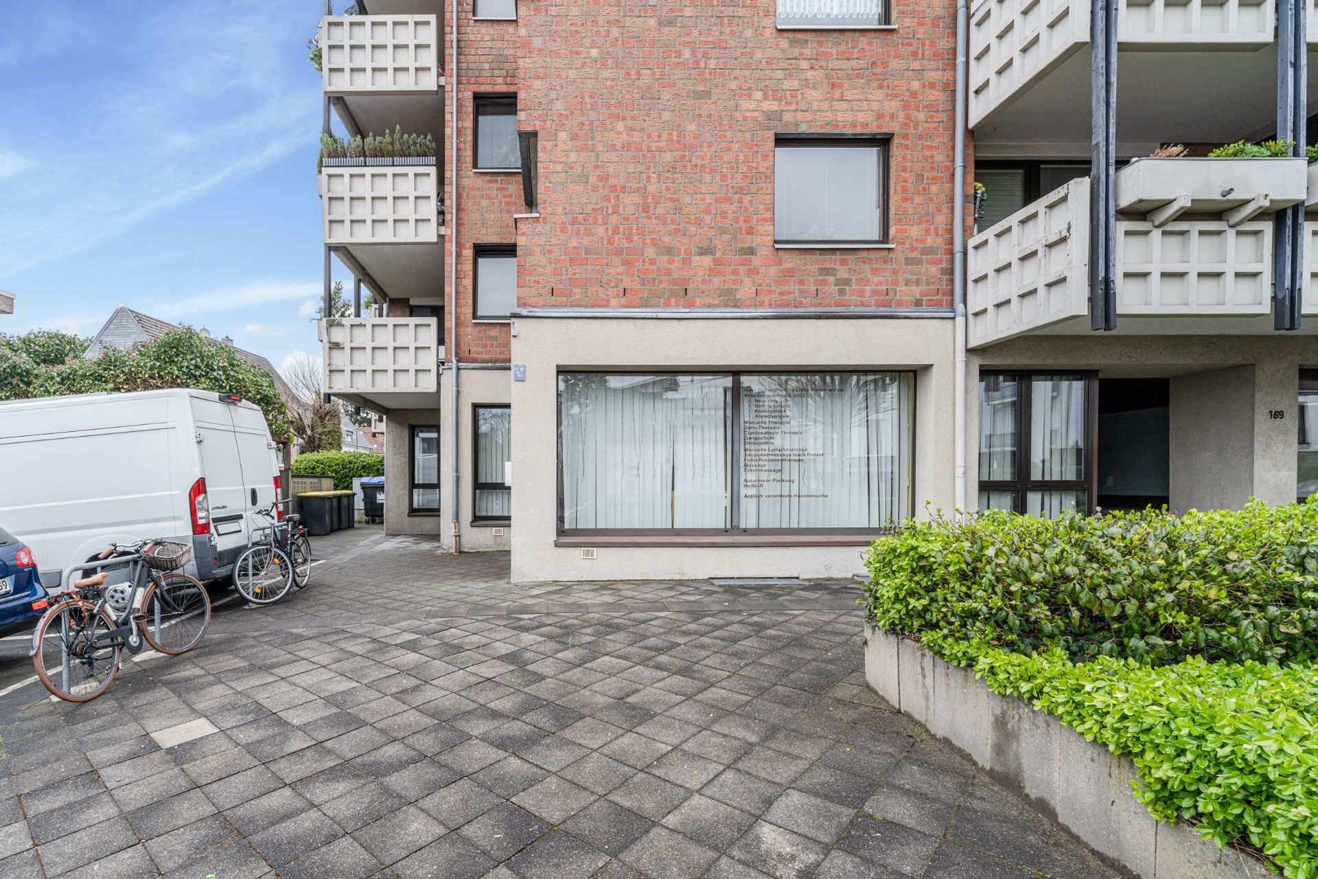 Mehrfamilienhaus mit Balkonen, großen Fenstern, geparktem Lieferwagen und Fahrrädern auf gepflastertem Vorplatz.
