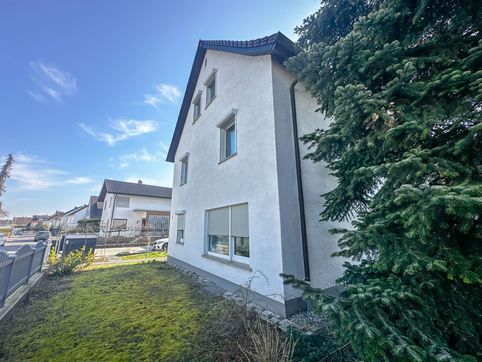 Weißes Wohnhaus mit mehreren Fenstern, Vorgarten und großen Nadelbaum im Vordergrund.