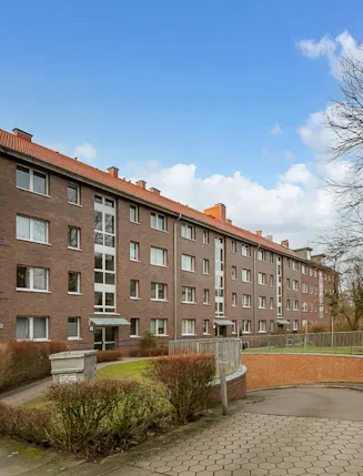 Viergeschossiges Mehrfamilienhaus aus Backstein mit Vorgarten und Gehweg bei Tageslicht.