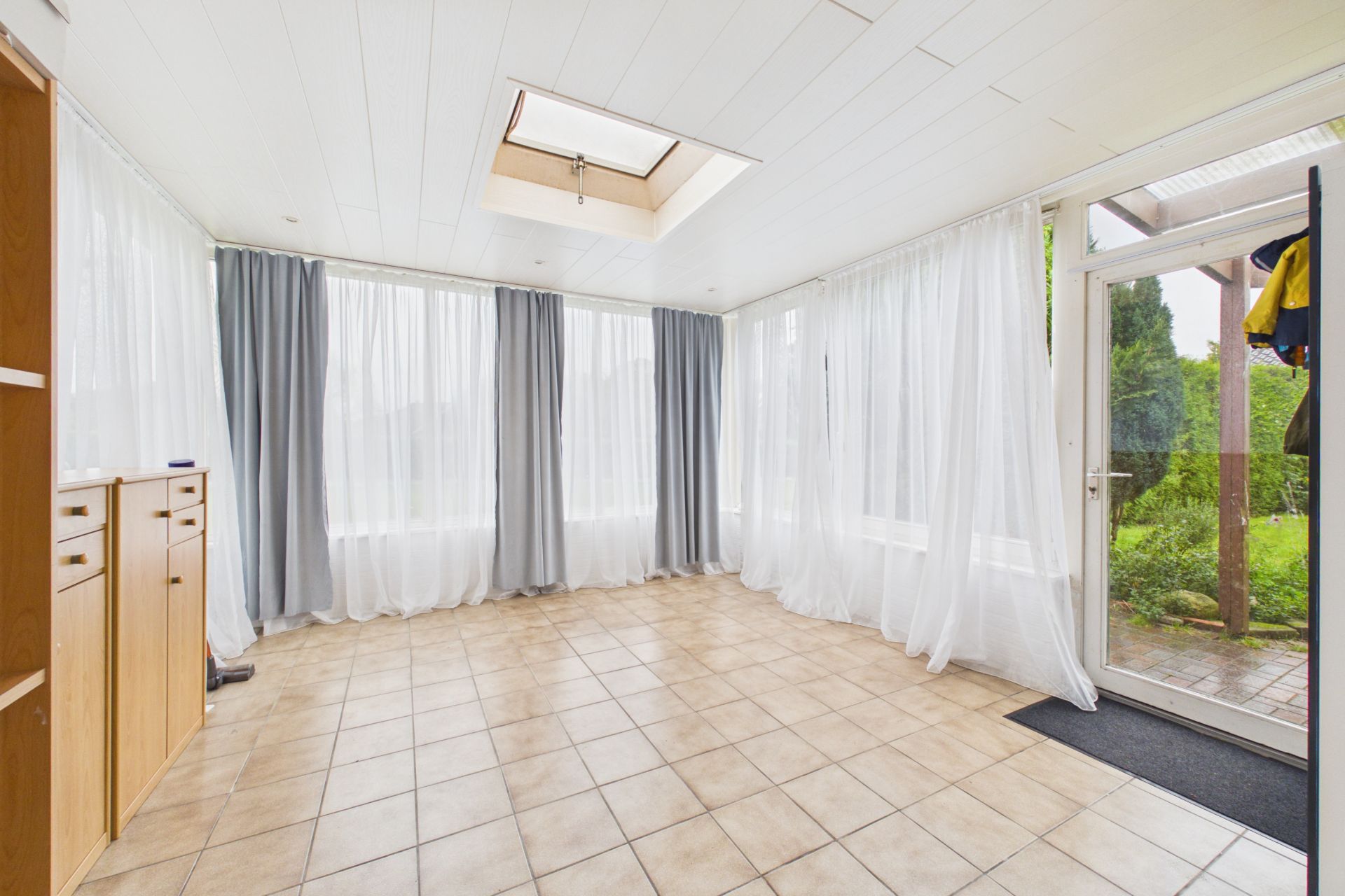 Heller Raum mit Fliesenboden, großen Fenstern, Vorhängen und Tür zum Garten.
