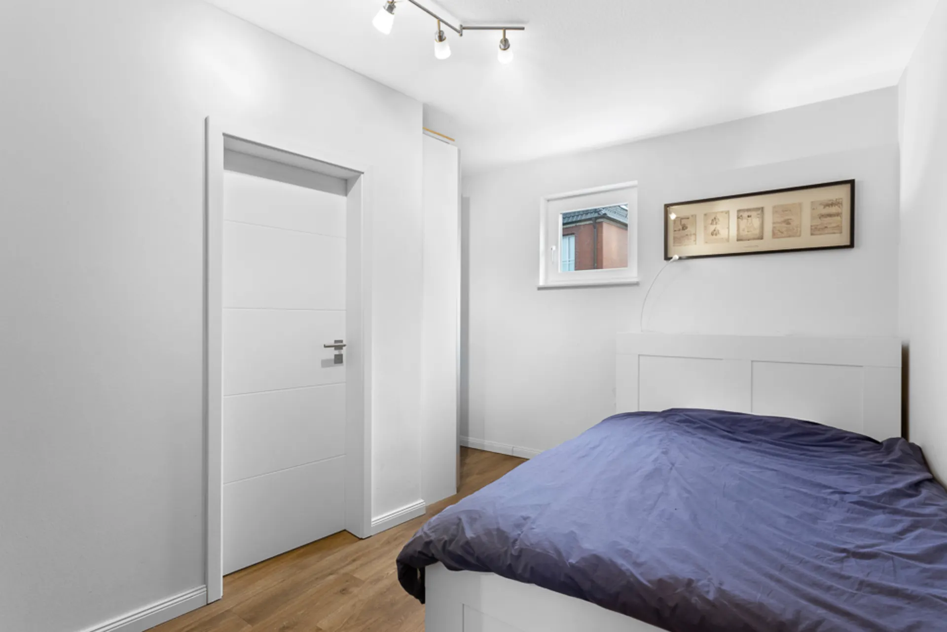Helles Schlafzimmer mit einem Bett, Holzboden, Fenster und weißer Tür.