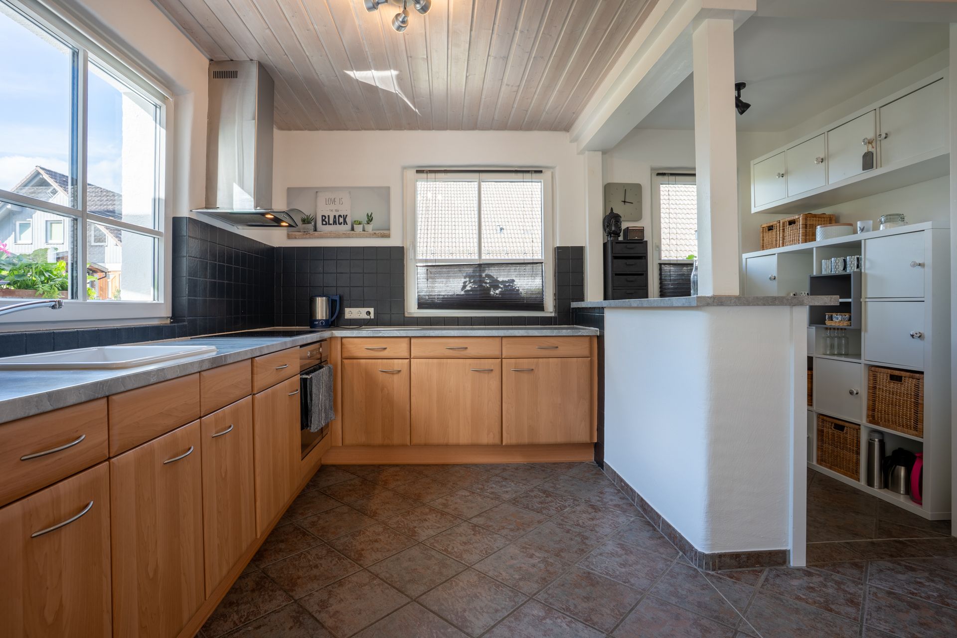 Moderne Küche mit Holzmöbeln, grauen Arbeitsplatten und großen Fenstern, daneben ein Regal mit Körben.