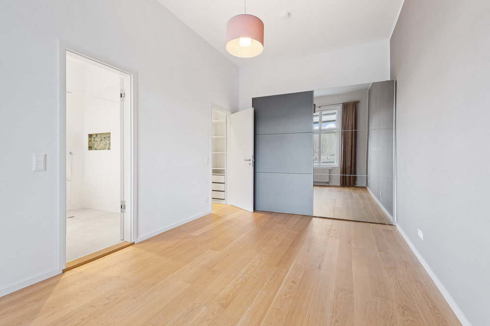 Helles Zimmer mit Holzfußboden, Spiegelwand, Schrank und Zugang zu Badezimmer und Nebenraum.