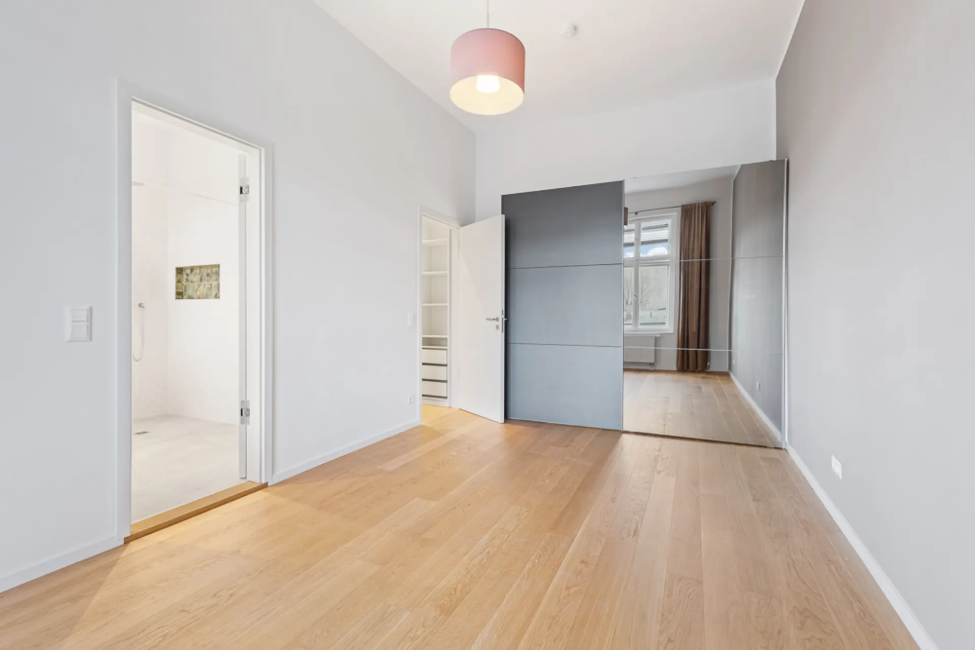 Helles Zimmer mit Holzfußboden, Spiegelwand, Schrank und Zugang zu Badezimmer und Nebenraum.