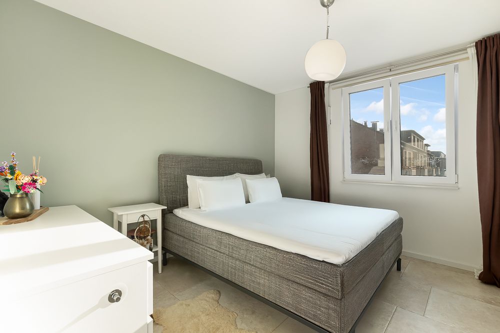 Helles Schlafzimmer mit Doppelbett, Nachttisch und Fenster mit braunen Vorhängen.