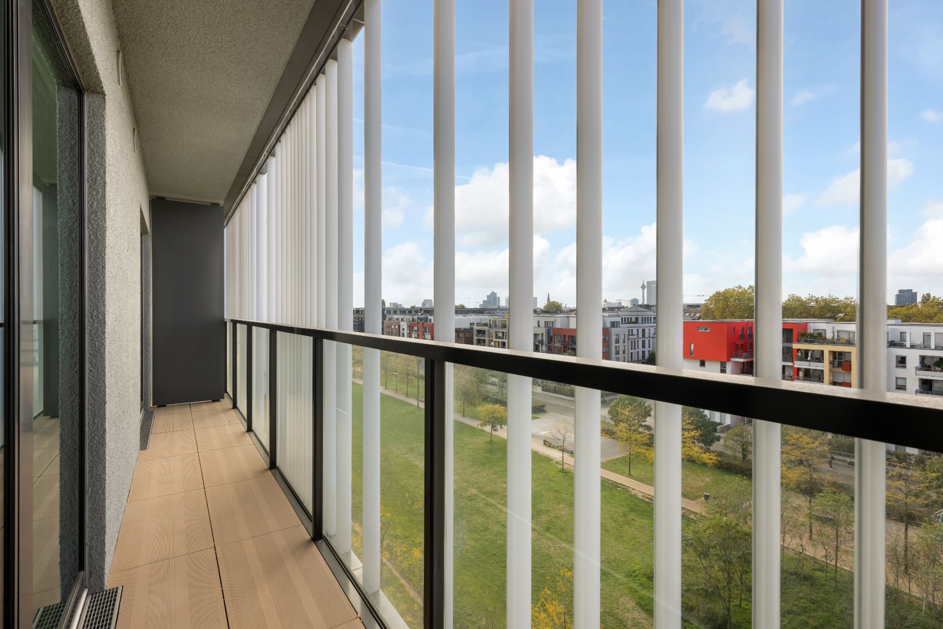 Balkon mit Glasgeländer und Blick auf einen grünen Innenhof und umliegende Wohnhäuser.