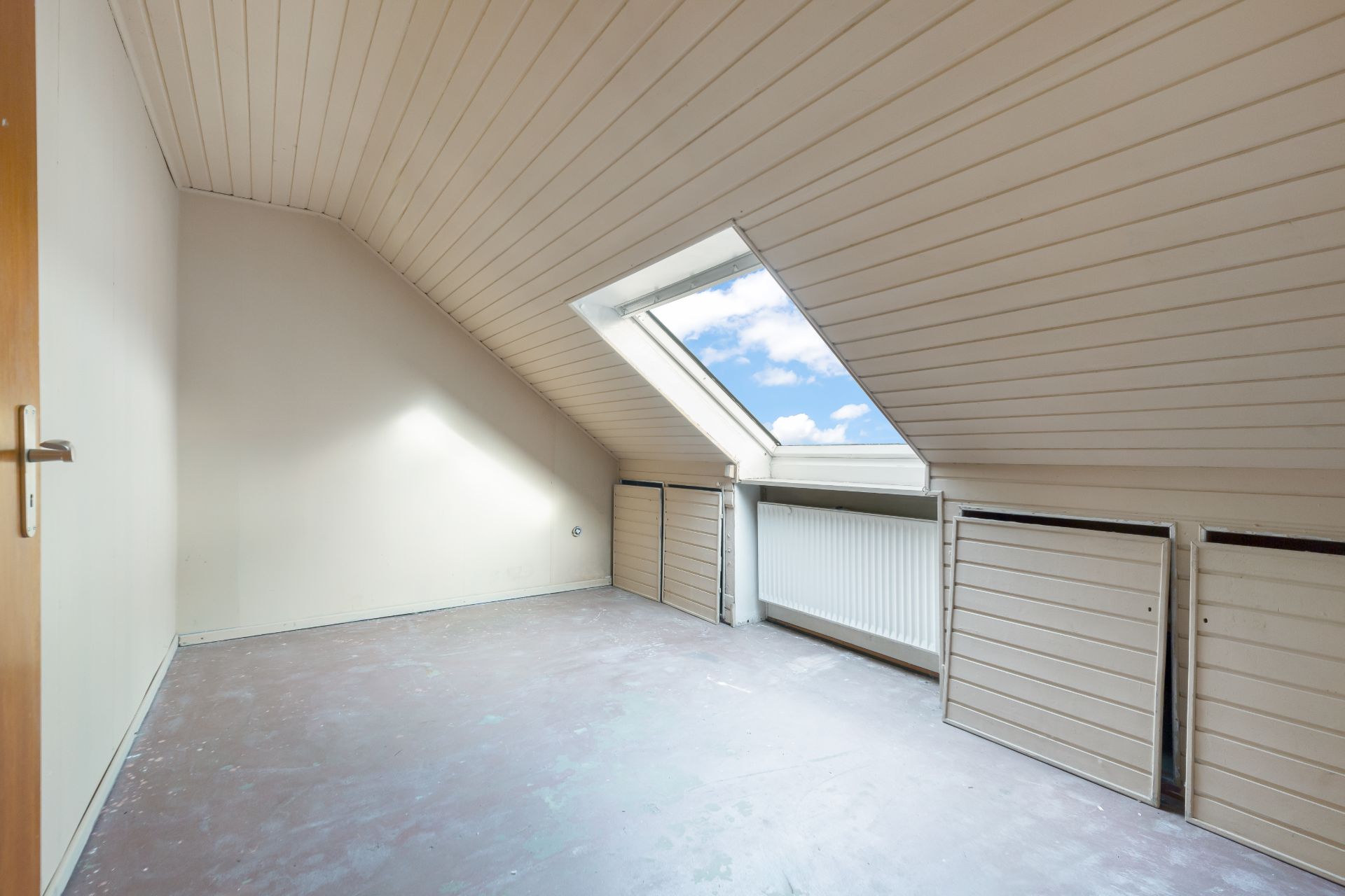 Leerer Dachgeschossraum mit Dachfenster, weißer Holzdecke und sichtbarem Heizkörper.