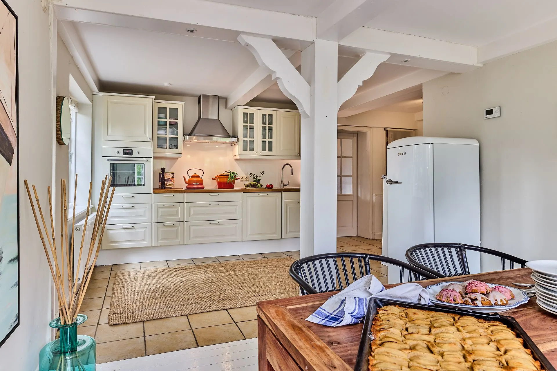 Helle Küche mit weißen Schränken, großem Kühlschrank und gedecktem Holztisch mit Gebäck.