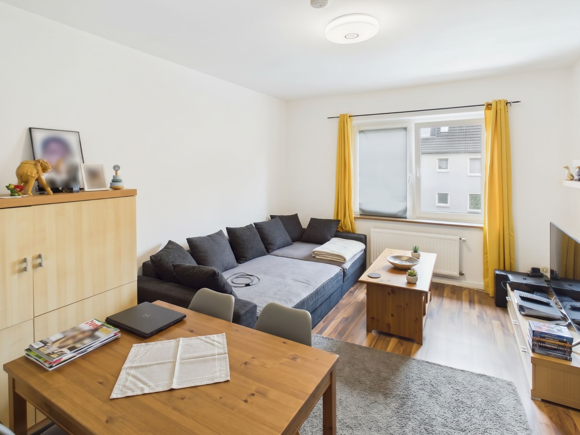 Wohnzimmer mit grauem Sofa, Holztisch, Esstisch, Fernseher und Fenster mit gelben Vorhängen.