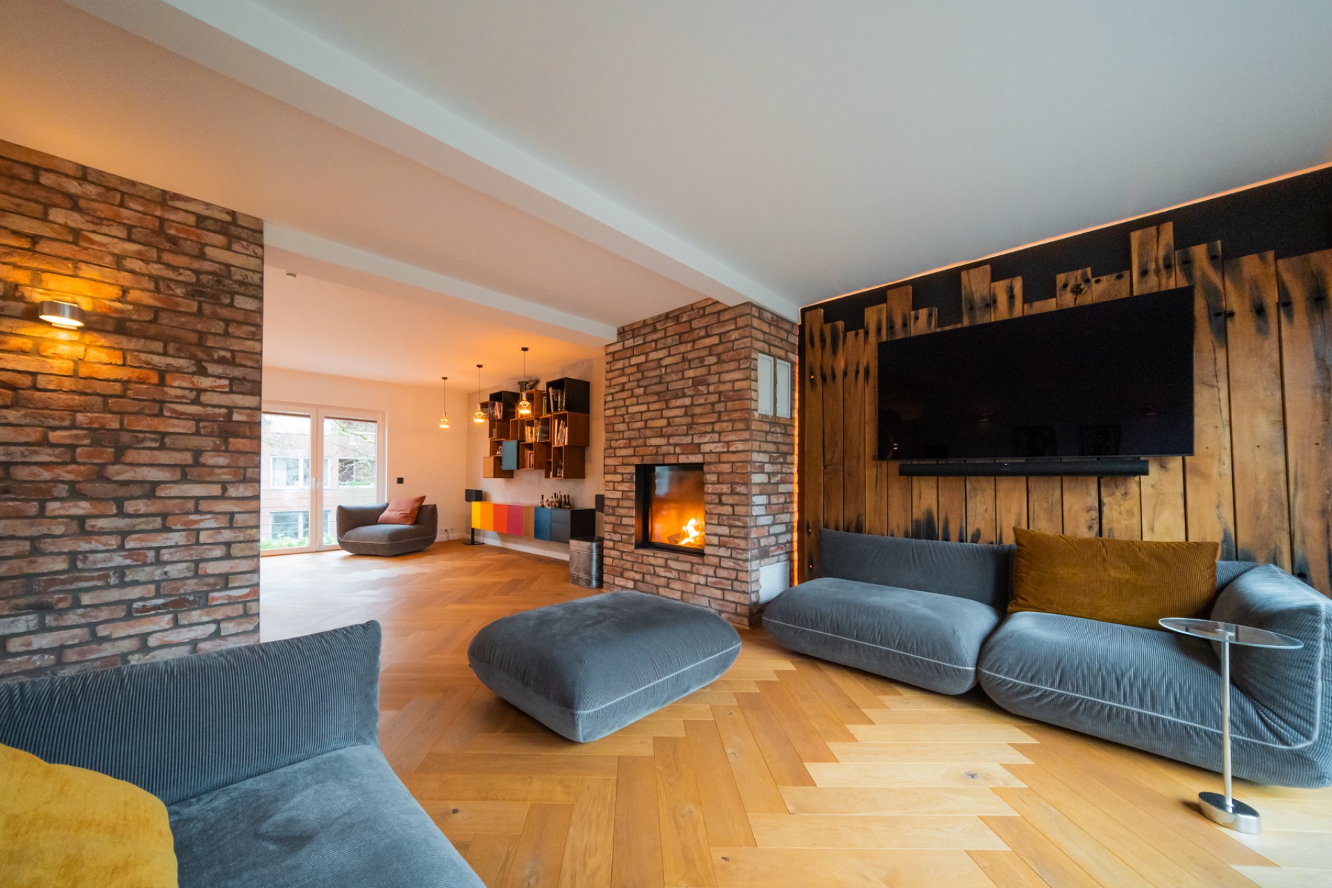 Modernes Wohnzimmer mit grauen Sofas, Kamin und Holzwand, daneben ein großer Fernseher.