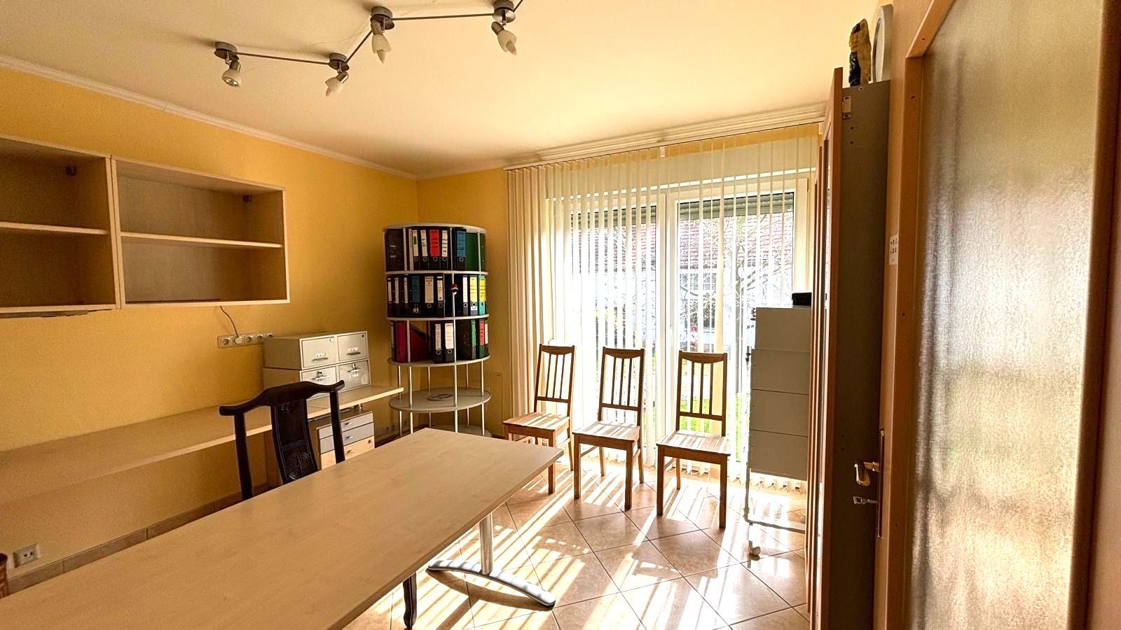 Helles Arbeitszimmer mit Tisch, Stühlen, Regalen und Fenstern mit Lamellenvorhängen.