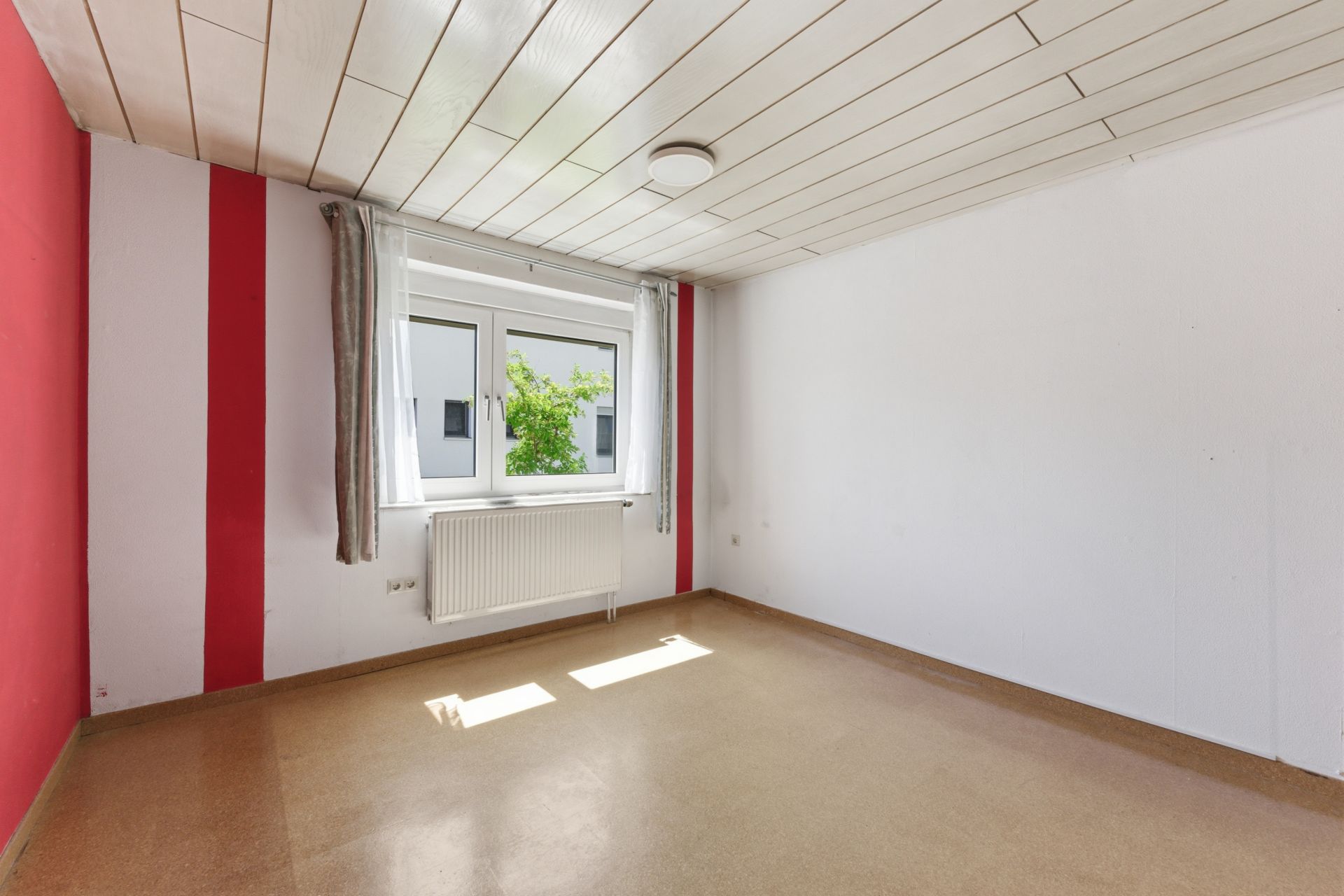 Leeres Zimmer mit weißer Decke, roten Akzenten, Fenster und Heizkörper.