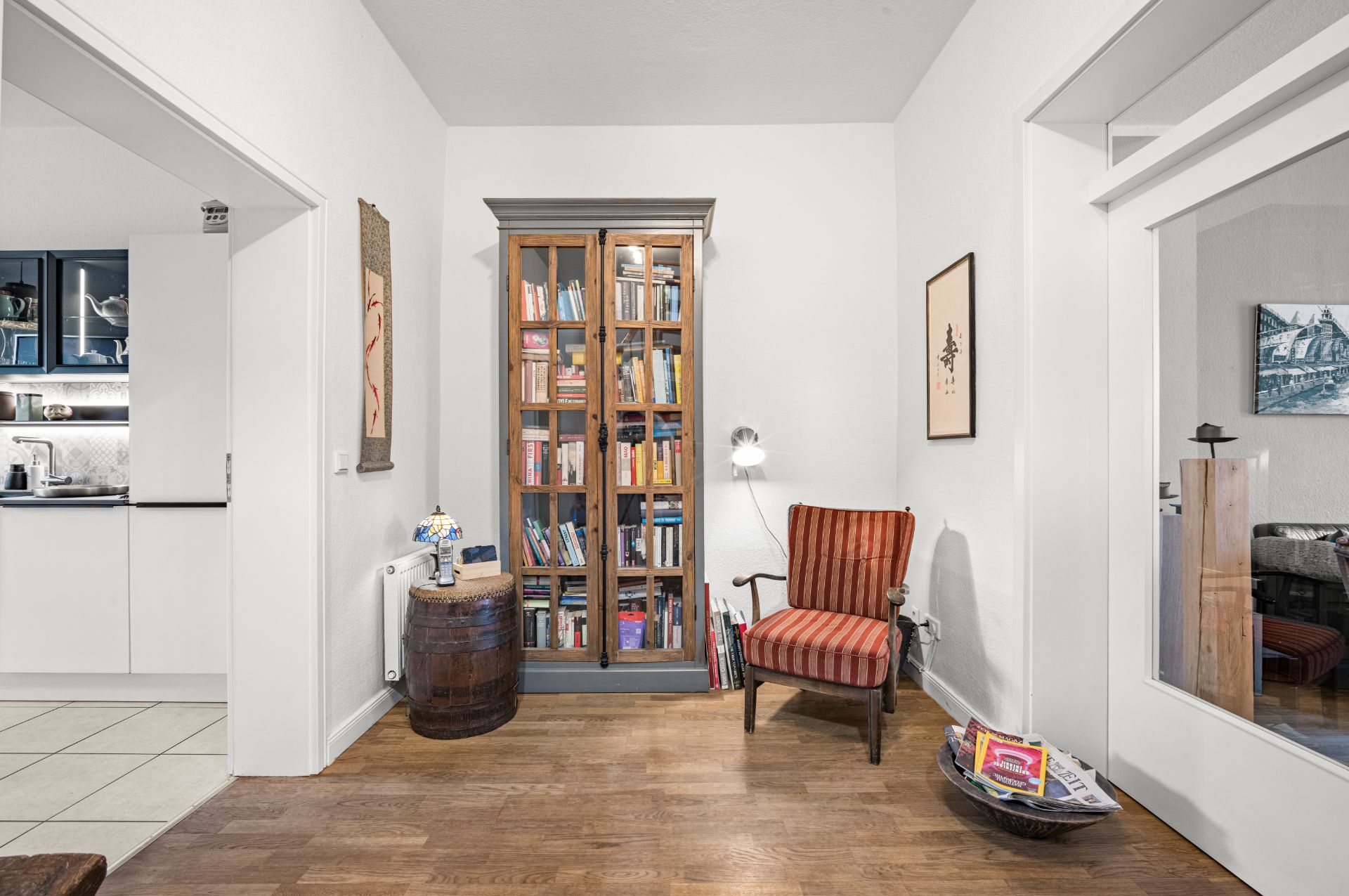 Wohnzimmer mit Bücherregal, Sessel, Stehlampe und Blick in eine Küche.