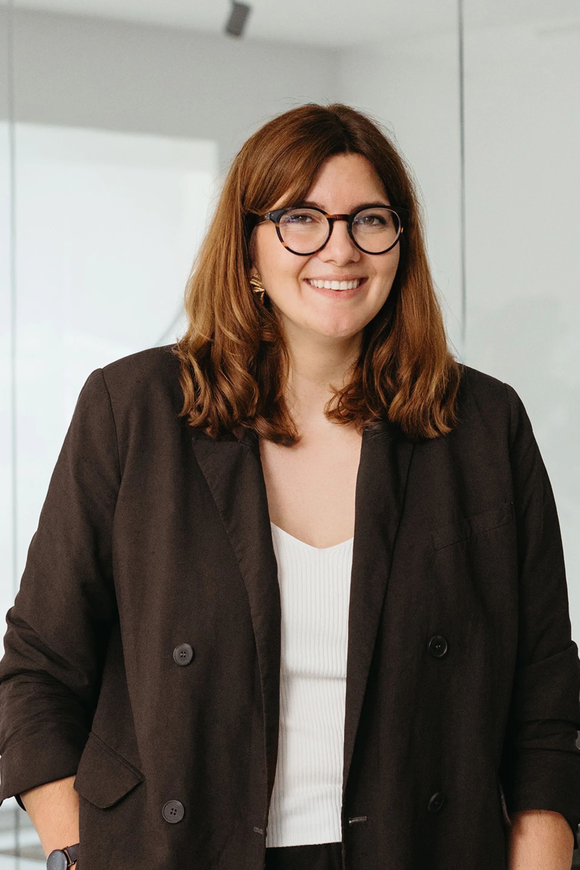Eine Frau mit Brille und brauner Jacke steht in einem hellen Raum mit Glaswand.