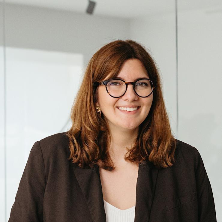 Eine Frau mit Brille und brauner Jacke steht in einem hellen Raum mit Glaswand.
