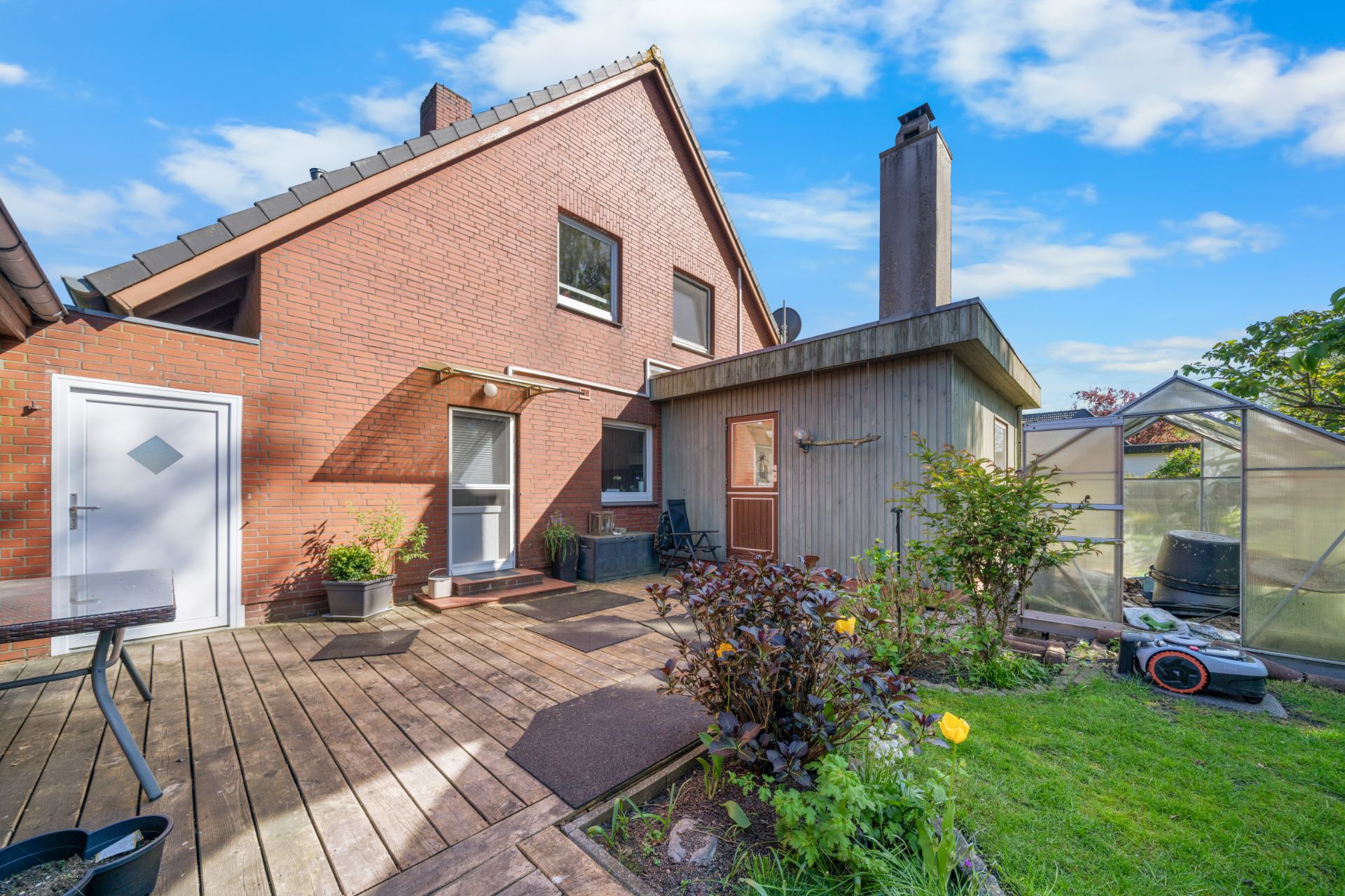 Hinterhof mit Terrasse, roten Ziegelhaus, Wintergarten und Garten mit Rasen und Pflanzen.