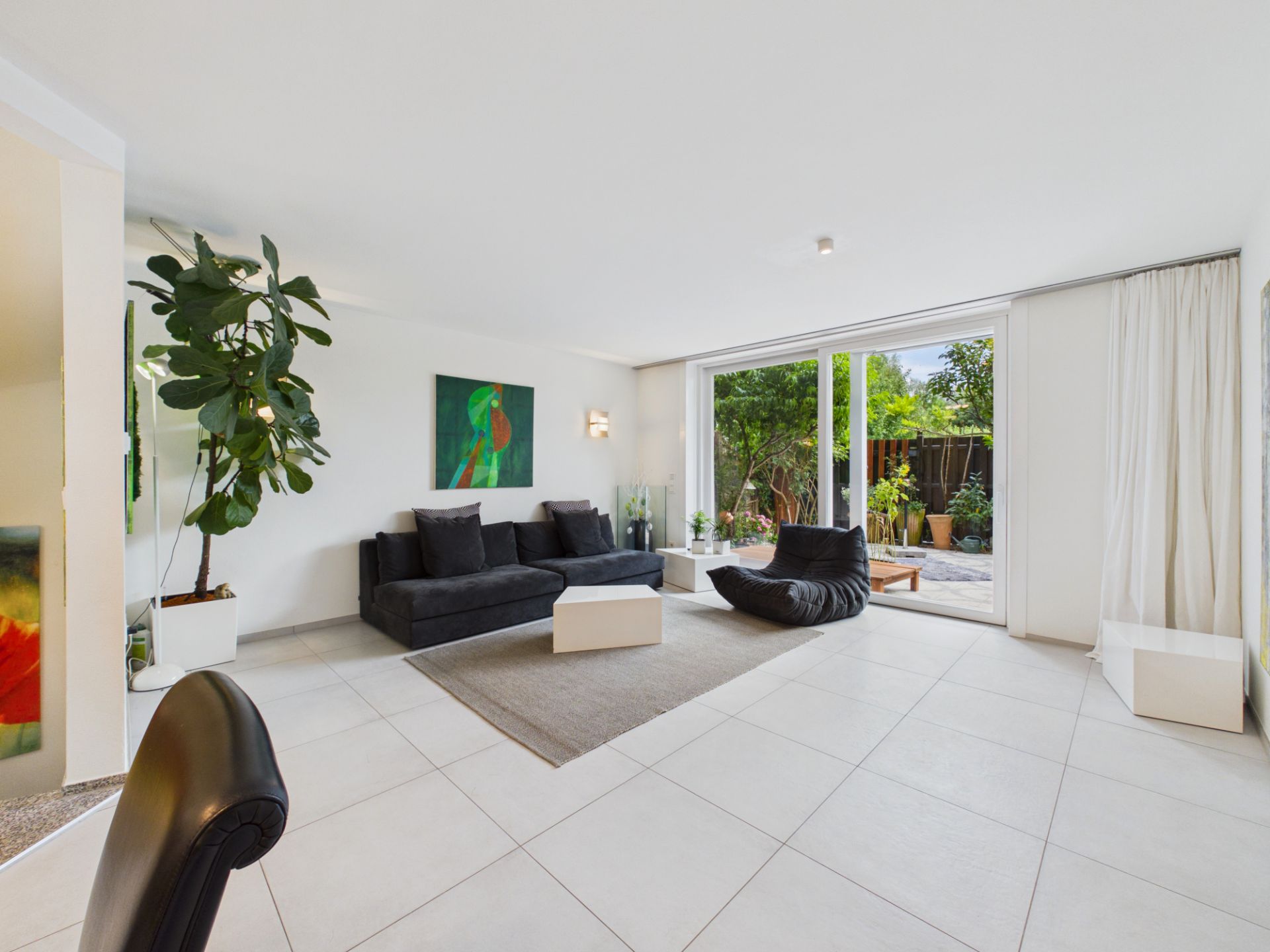Helles Wohnzimmer mit schwarzem Sofa, großer Pflanze und Zugang zur Terrasse.
