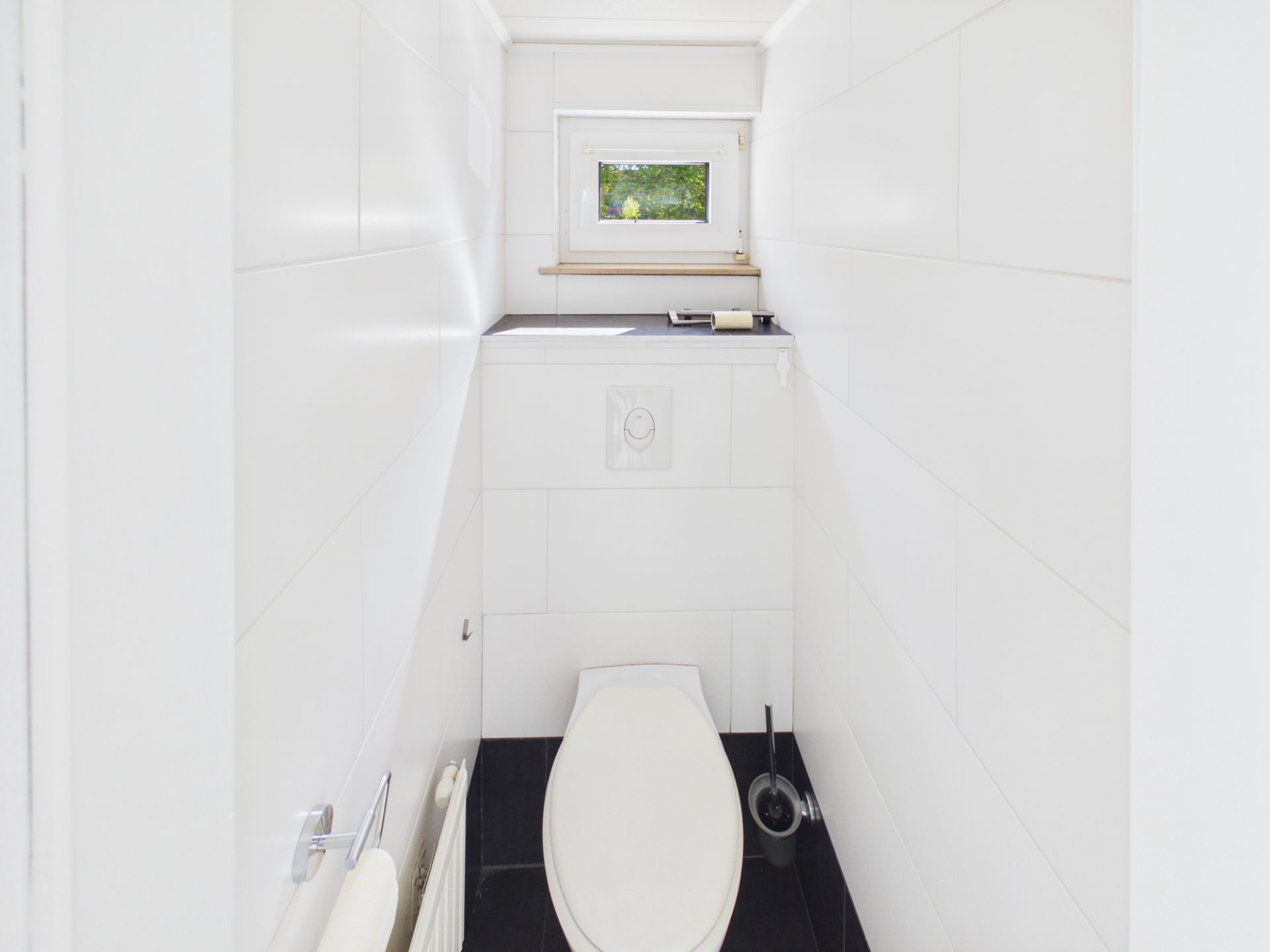 Kleiner, weiß gefliester Toilettenraum mit WC, Toilettenbürste und kleinem Fenster.