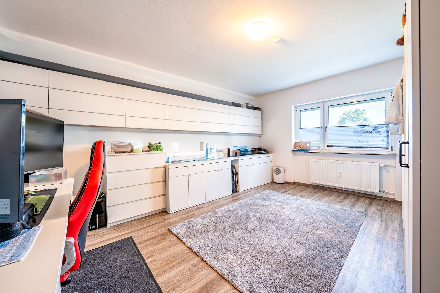 Helles Arbeitszimmer mit Schreibtisch, rotem Stuhl, Regalen und großem Fenster.