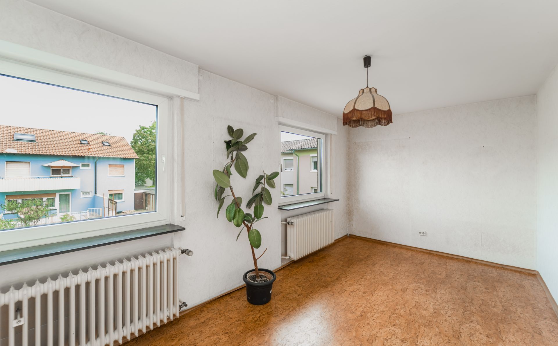 Helles Zimmer mit zwei Fenstern, Heizkörper, einer Topfpflanze und Hängelampe, Sicht auf Nachbarhäuser.