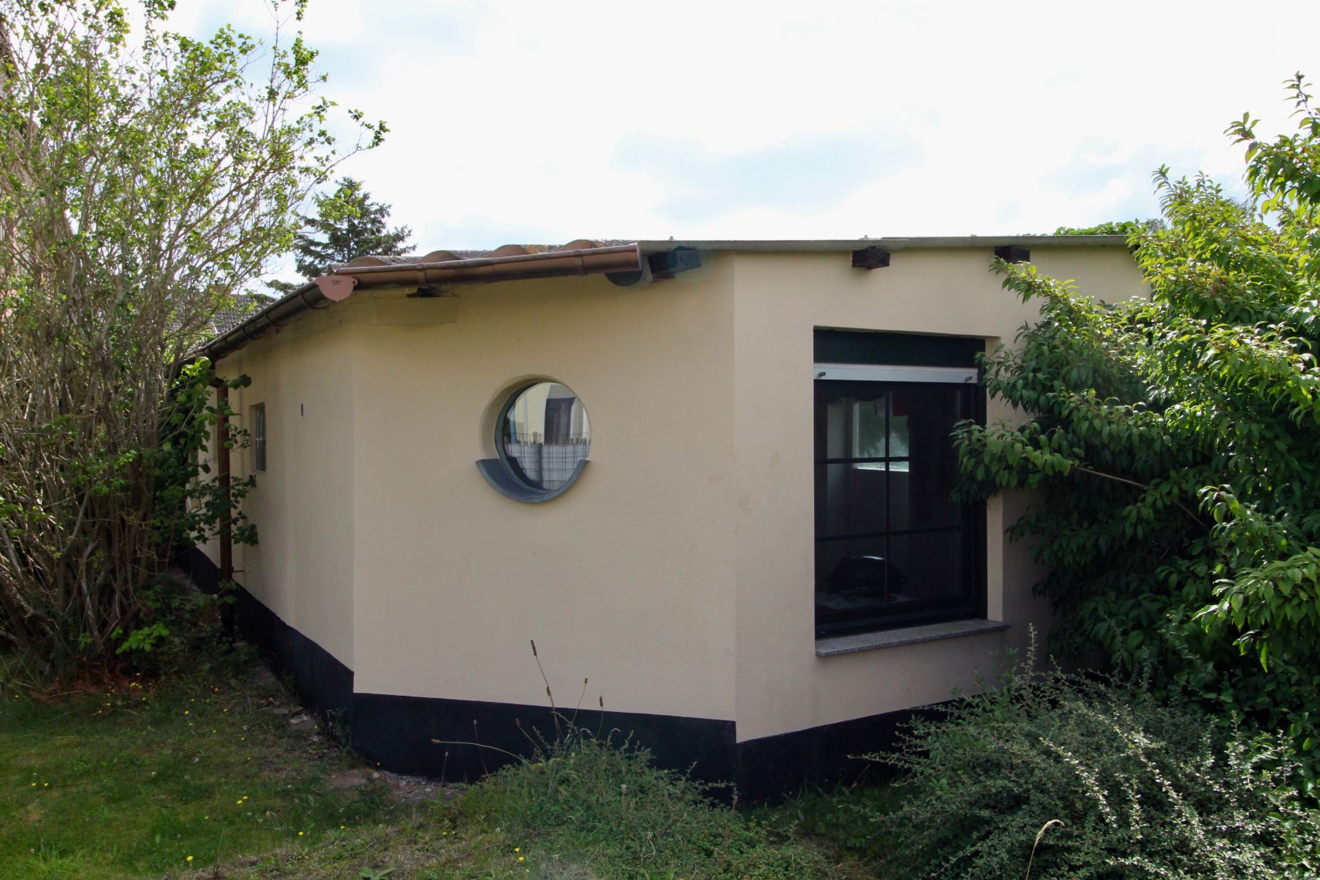 Eckiges, einstöckiges Haus mit rundem und eckigem Fenster, umgeben von Büschen.