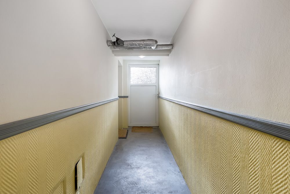 Langer Flur mit gelben Wänden, grauem Boden und einer weißen Tür am Ende.