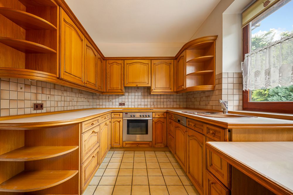 Große Einbauküche mit Holzfronten, Herd, Spüle und Eckregalen, Fliesenboden und Fenster.