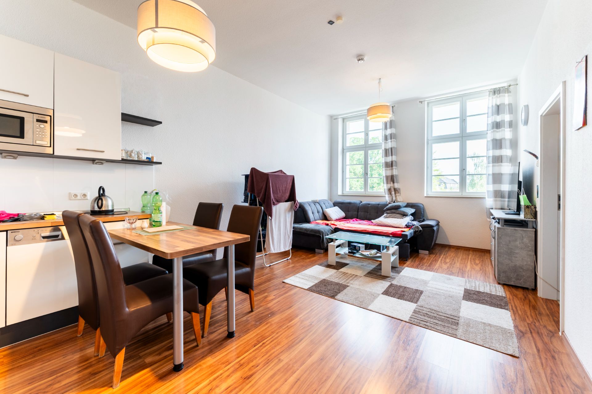Helles Wohnzimmer mit Ecksofa, Esstisch, offener Küche und großen Fenstern.