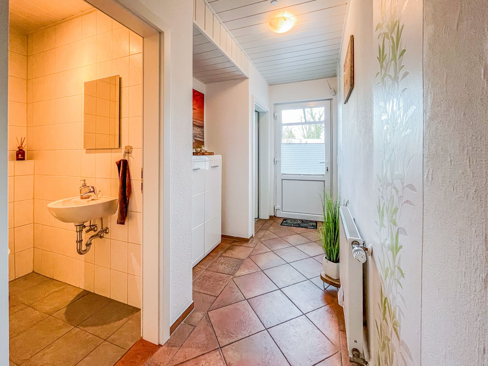 Heller Flur mit Heizung, Topfpflanze und offenem Zugang zu einem Badezimmer mit Waschbecken und Spiegel.