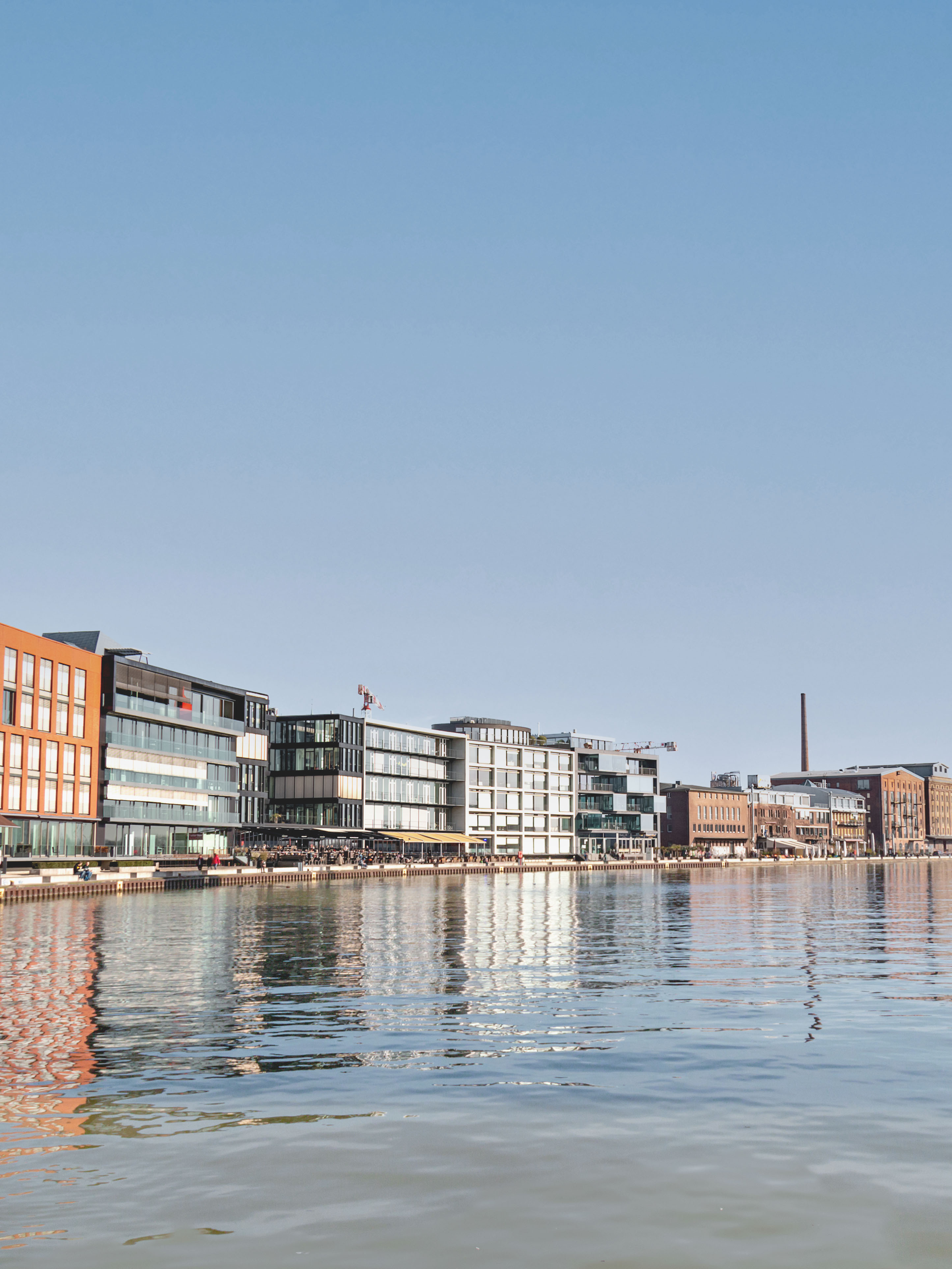 Moderne Wohngebäude und Promenade an einem ruhigen Flussufer bei sonnigem Wetter.
