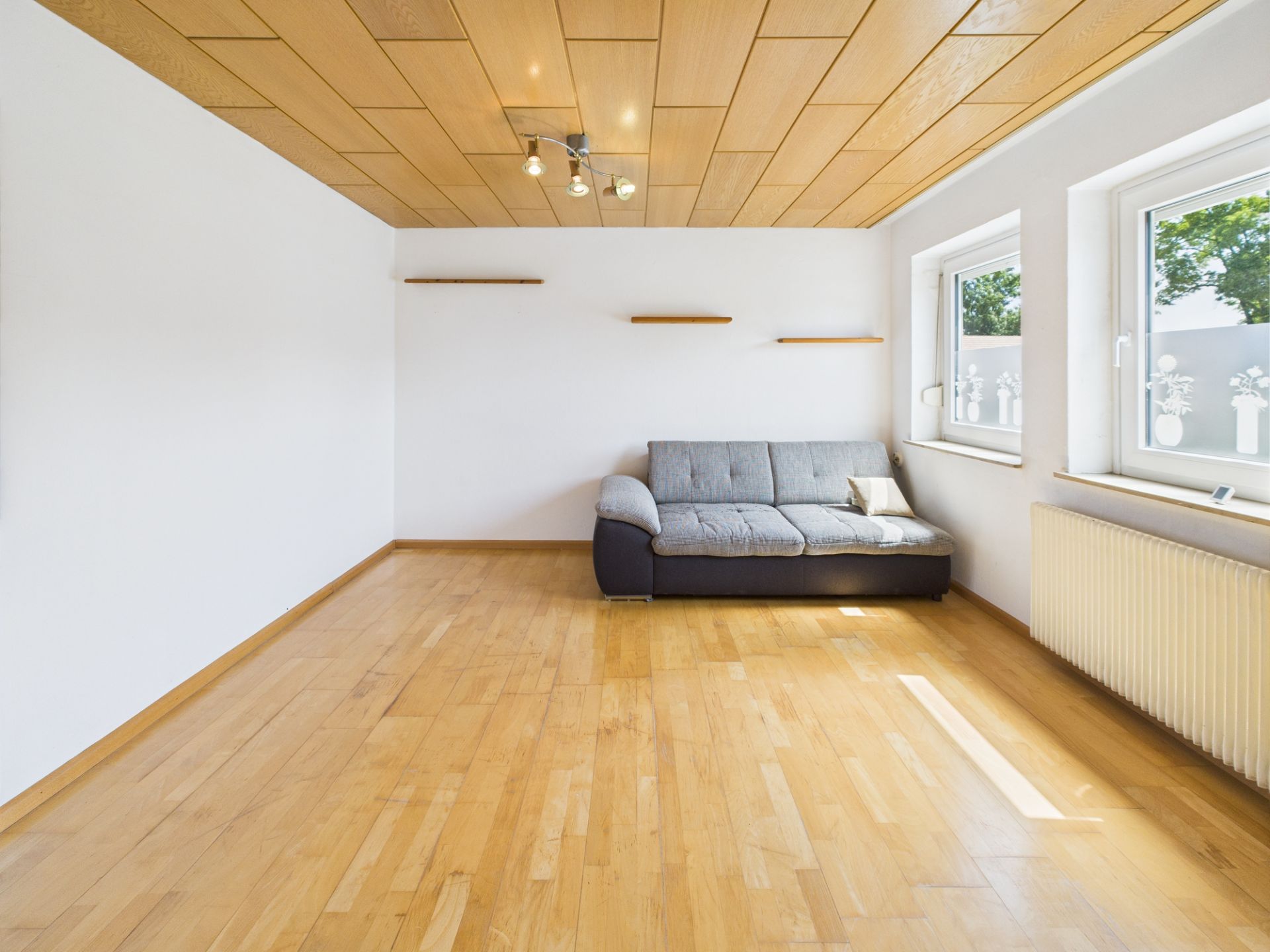Helles Wohnzimmer mit Holzfußboden, grauem Sofa und zwei Fenstern.