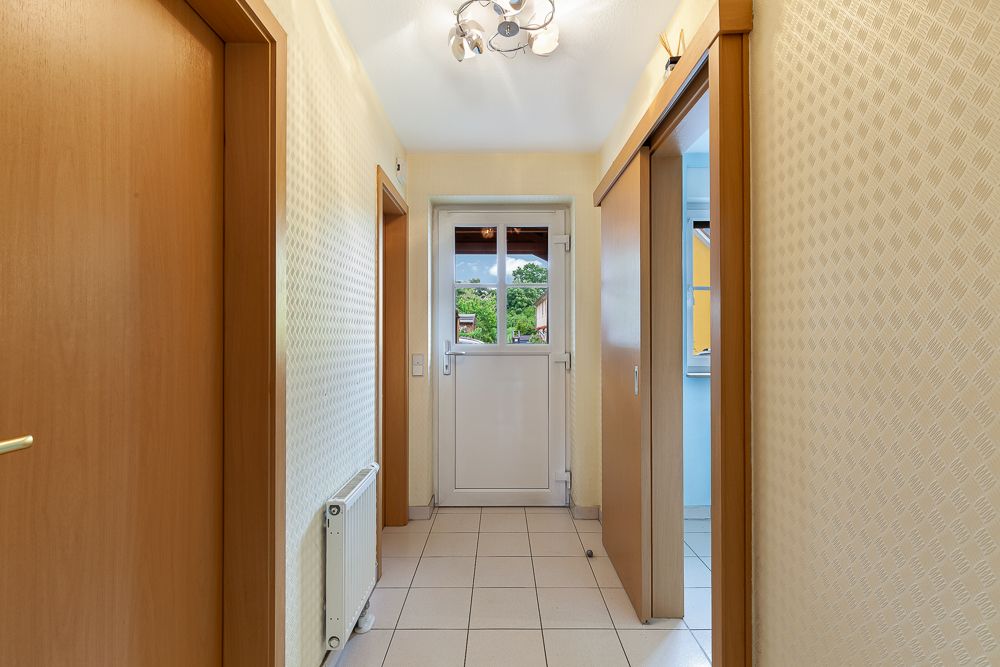 Heller Flur mit Fliesenboden, weißen Wänden und Holztüren, Tür mit Fenster am Ende.
