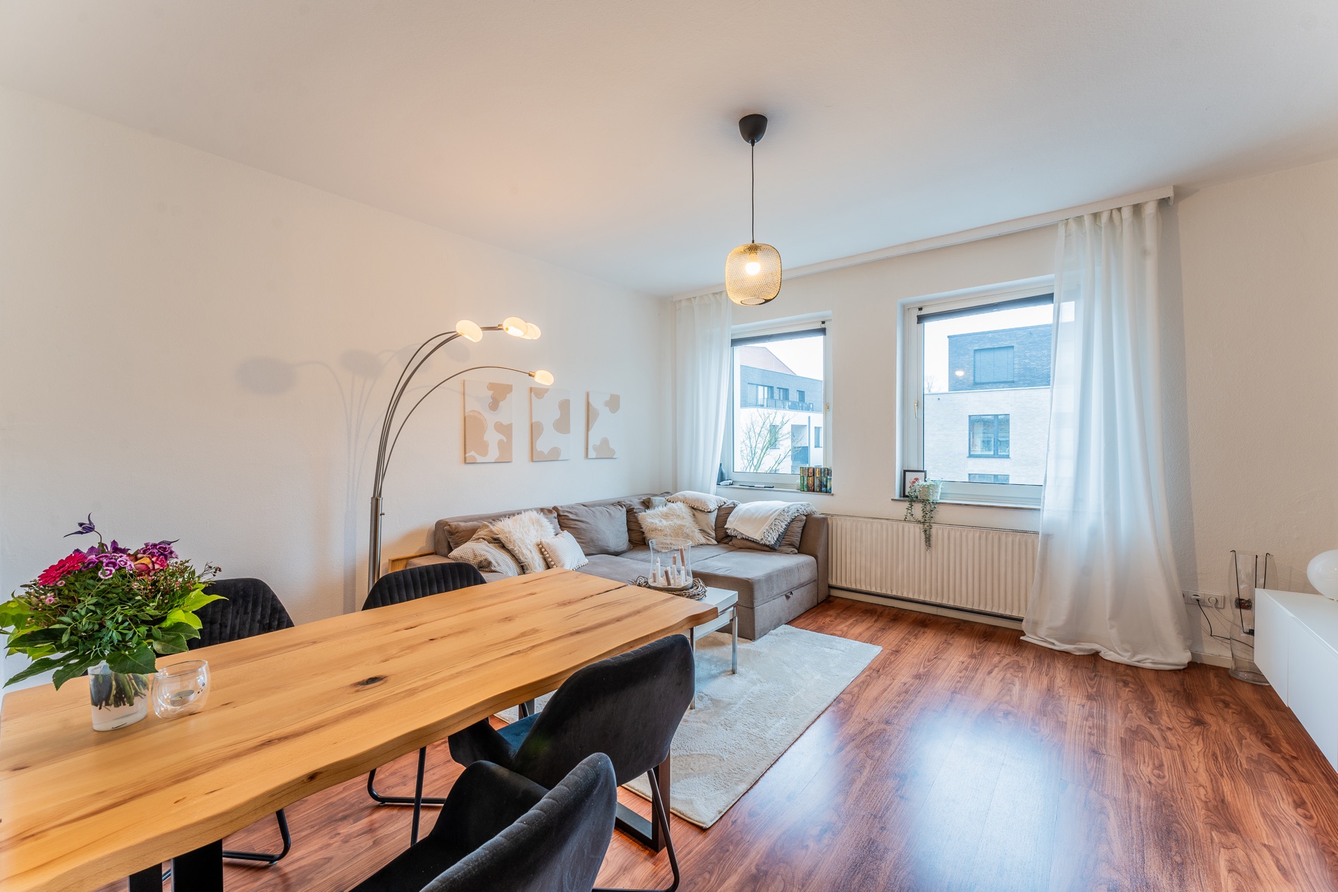 Helles Wohnzimmer mit Holztisch, Stühlen, Ecksofa, Stehlampe, Teppich und zwei Fenstern.