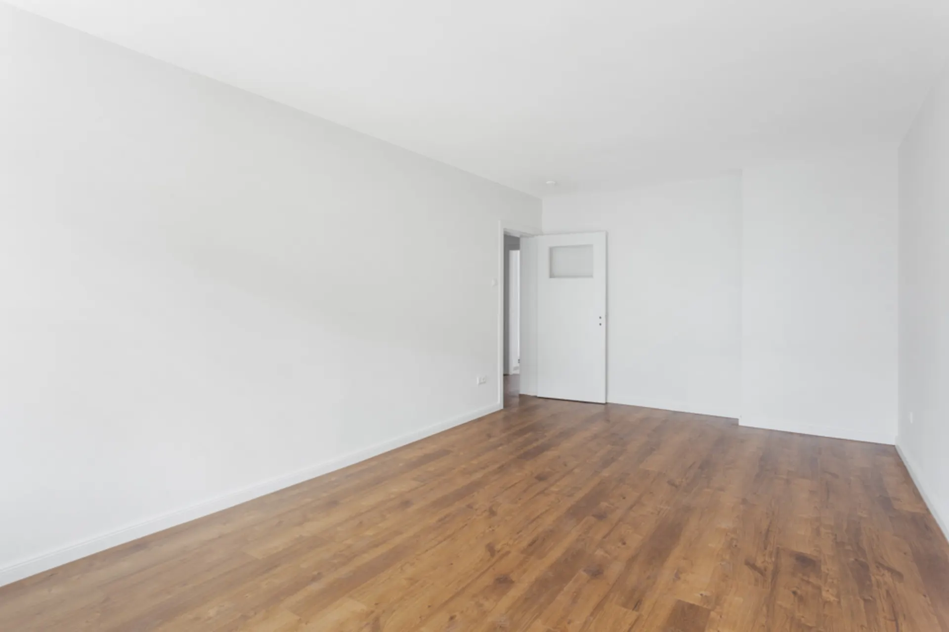 Leerer Raum mit weiß gestrichenen Wänden und braunem Holzboden, weiße Tür geöffnet.