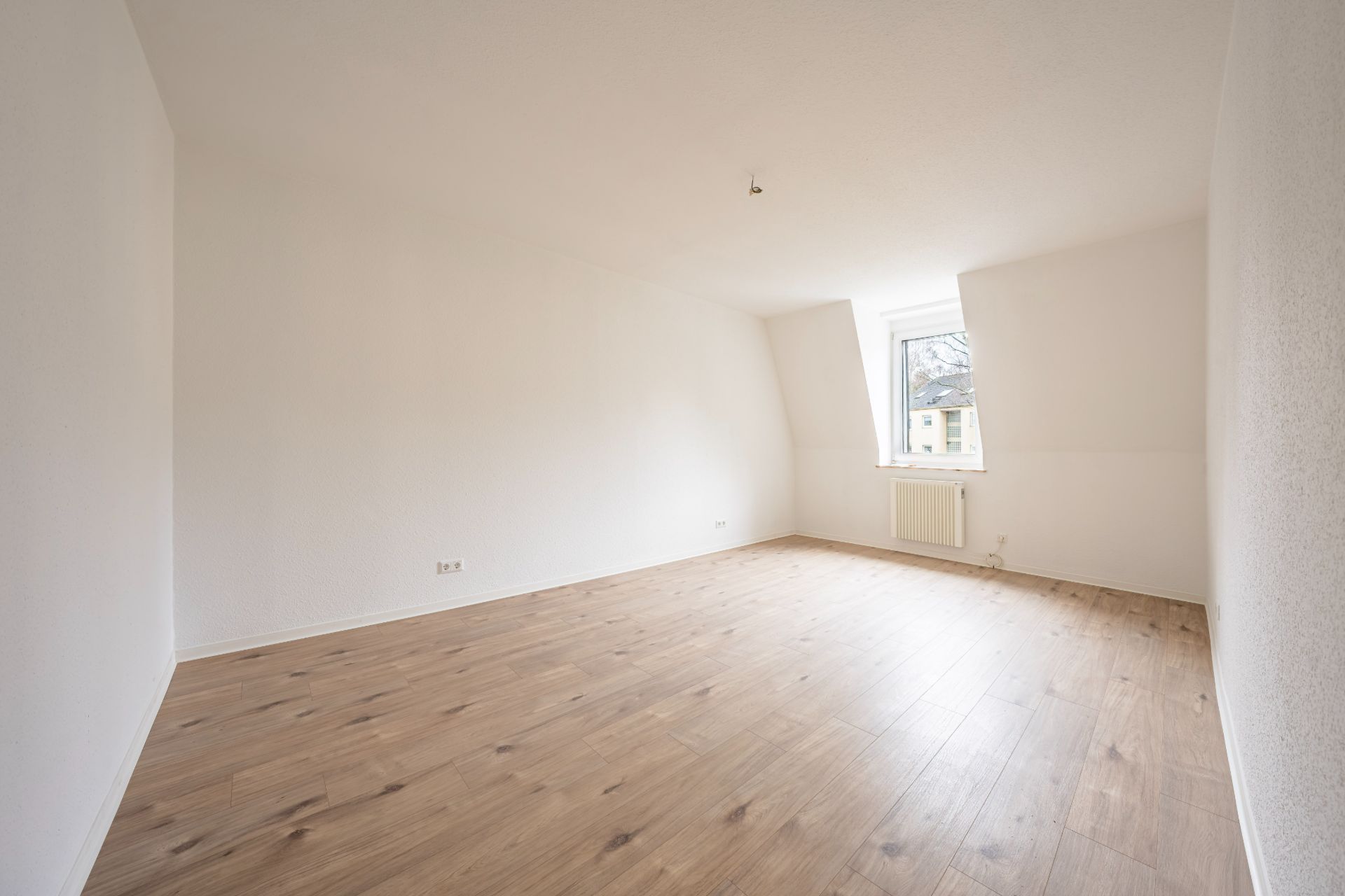 Leerer Raum mit weiß gestrichenen Wänden, Holzfußboden und einem Fenster mit Heizung darunter.