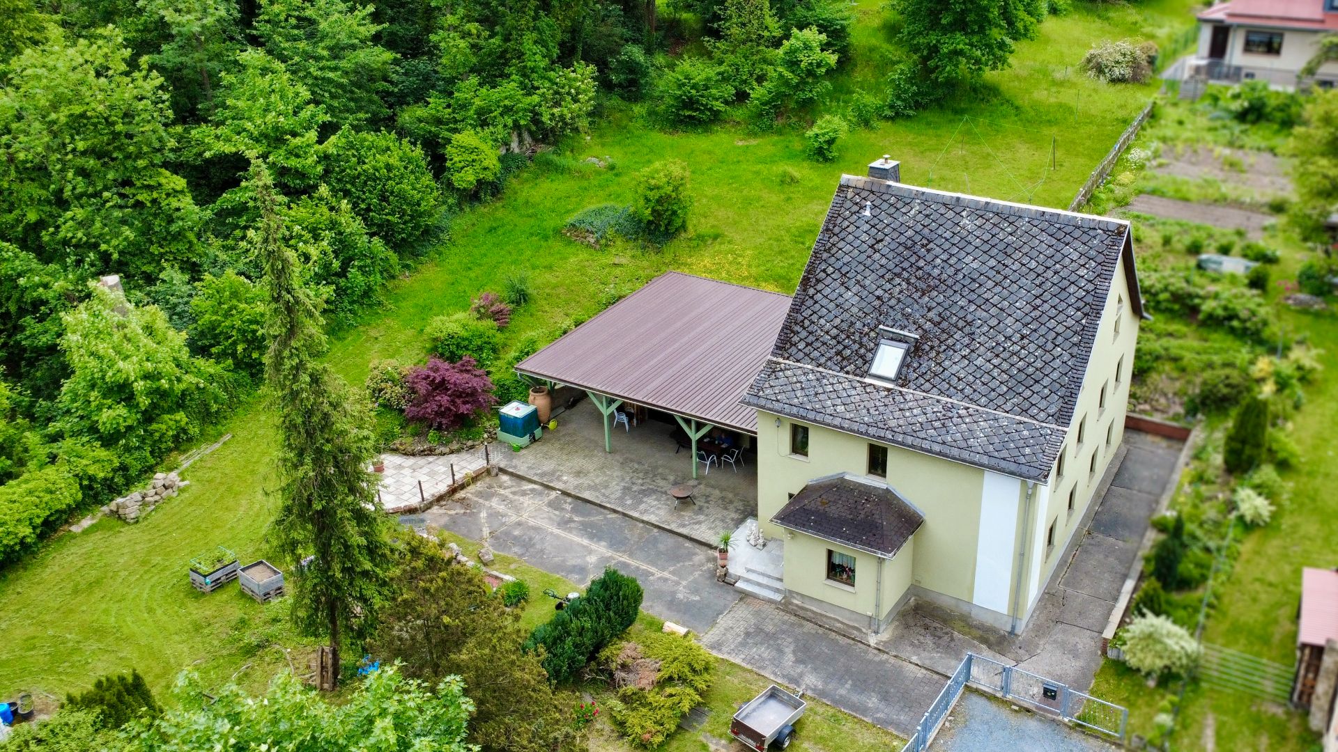 Haus mit überdachter Terrasse und großem Garten, von Bäumen und Grünflächen umgeben.